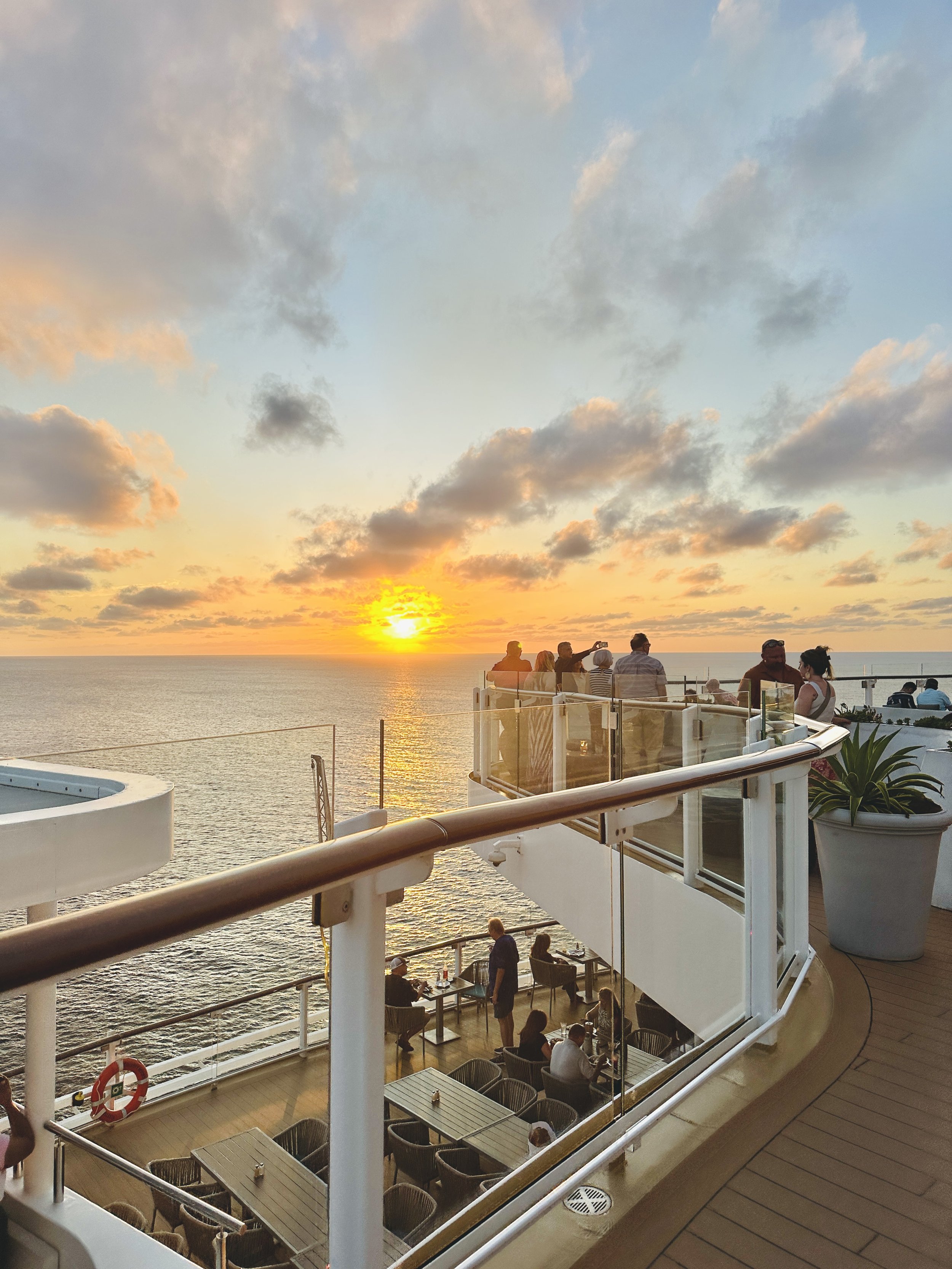 People dining and socializing on a cruise ship deck during sunset over the ocean with a partly cloudy sky.