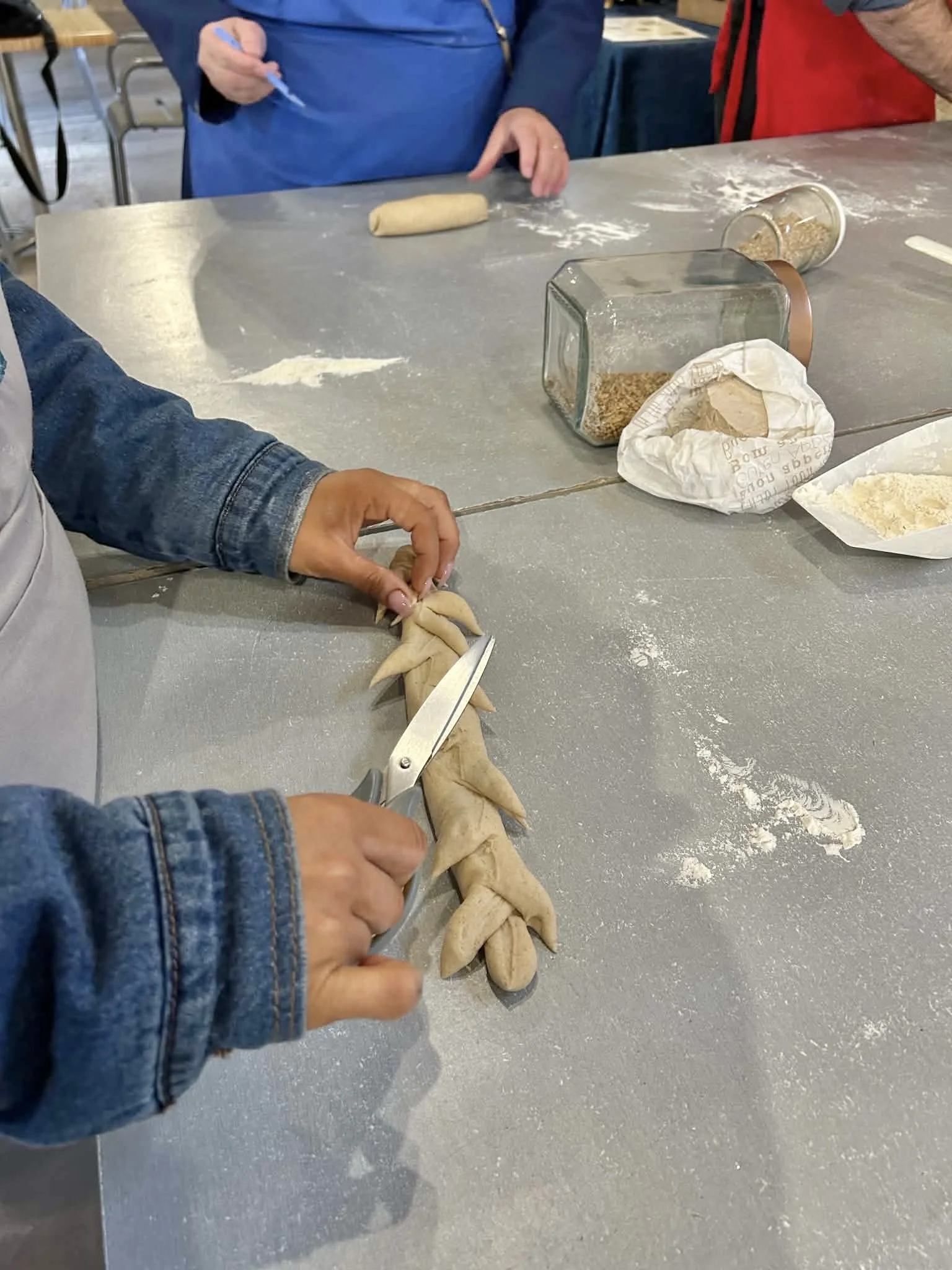 People shaping and cutting dough on a gray countertop, with flour, grains, and utensils nearby.