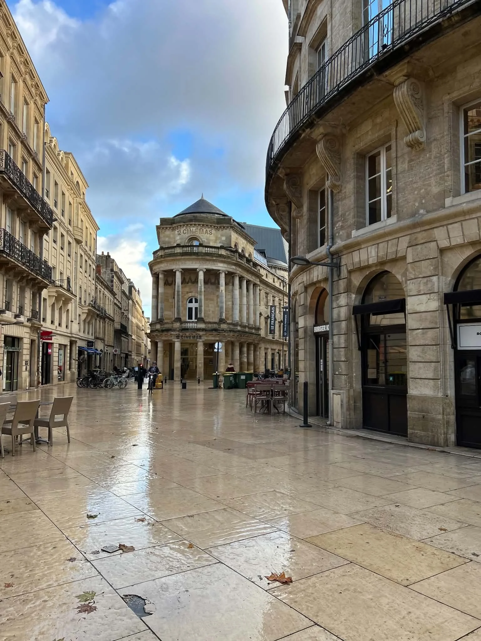 Empty city street with wet pavement, some fallen leaves, historic European-style buildings, bicycles, and a cinema sign visible.
