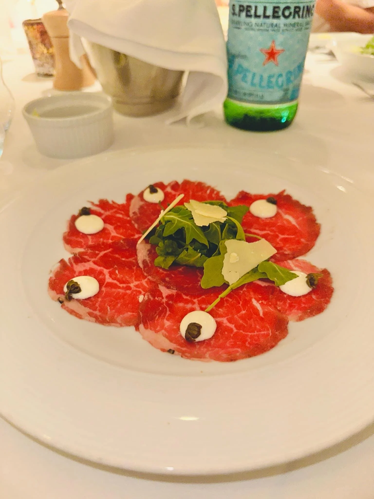 Thinly sliced raw beef carpaccio garnished with small dollops of white sauce, black peppercorns, fresh greens, and shaved cheese, served on a white plate at a dining table.