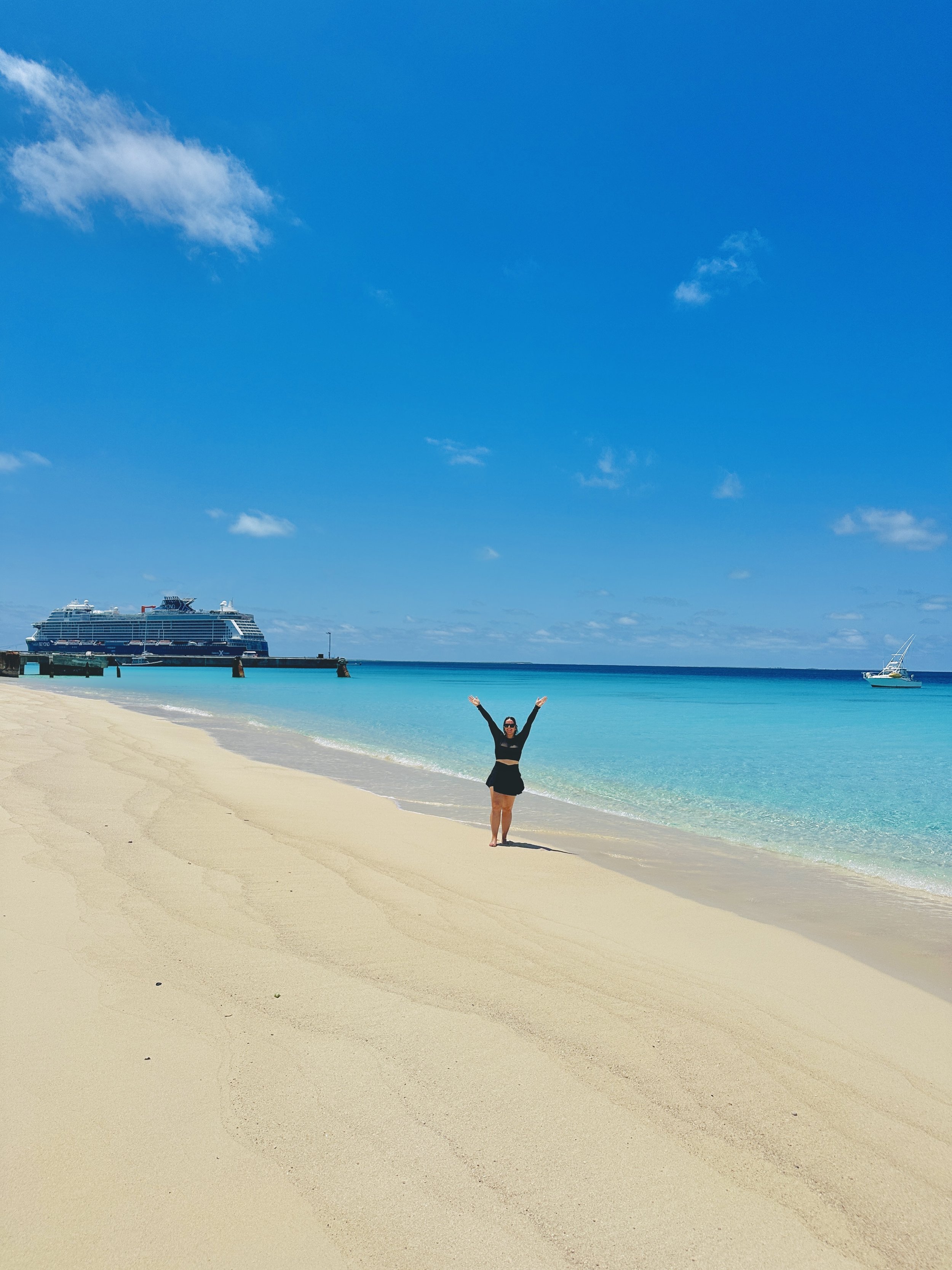 A woman in black swimsuit standing on a sandy beach with arms raised, clear blue sky, turquoise water, and a cruise ship and sailboat in the background.