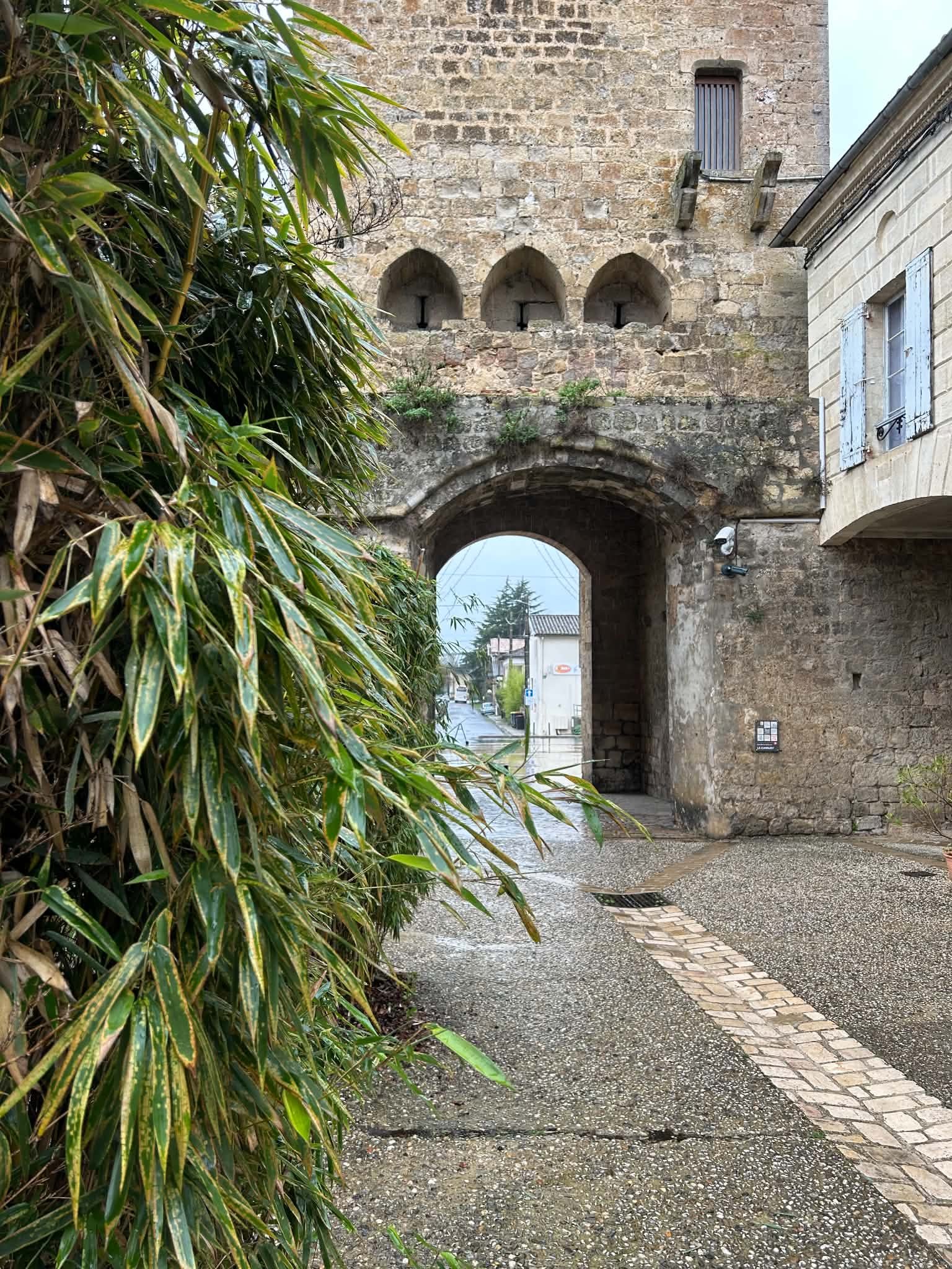 A stone archway leading to a street, with a stone building and greenery on the left and a building with blue shutters on the right.