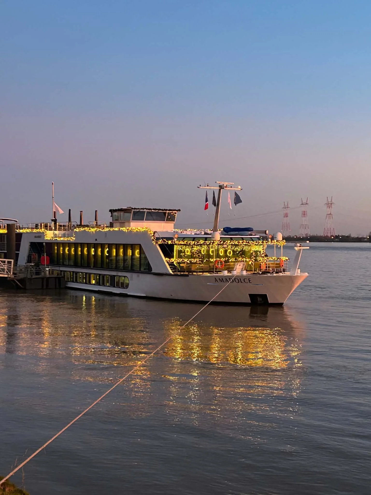 A cruise ship named Amalfi docked at a harbor during sunset, with lights illuminating the ship and antennas flying flags, and power lines in the background.