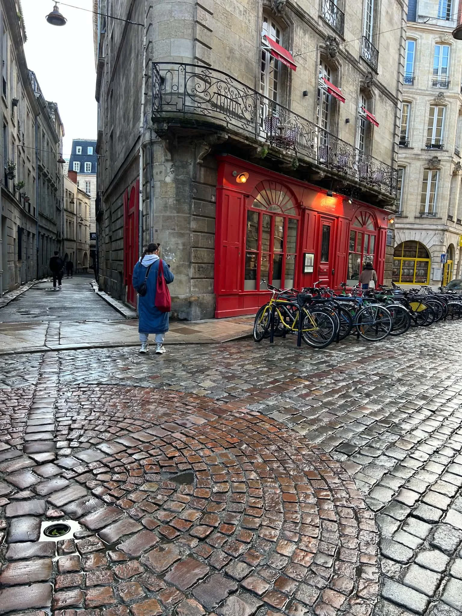 A cobblestone street corner in a European city with wet stones, a red storefront with large windows, bicycles parked in front, and old stone buildings with balconies and red awnings.
