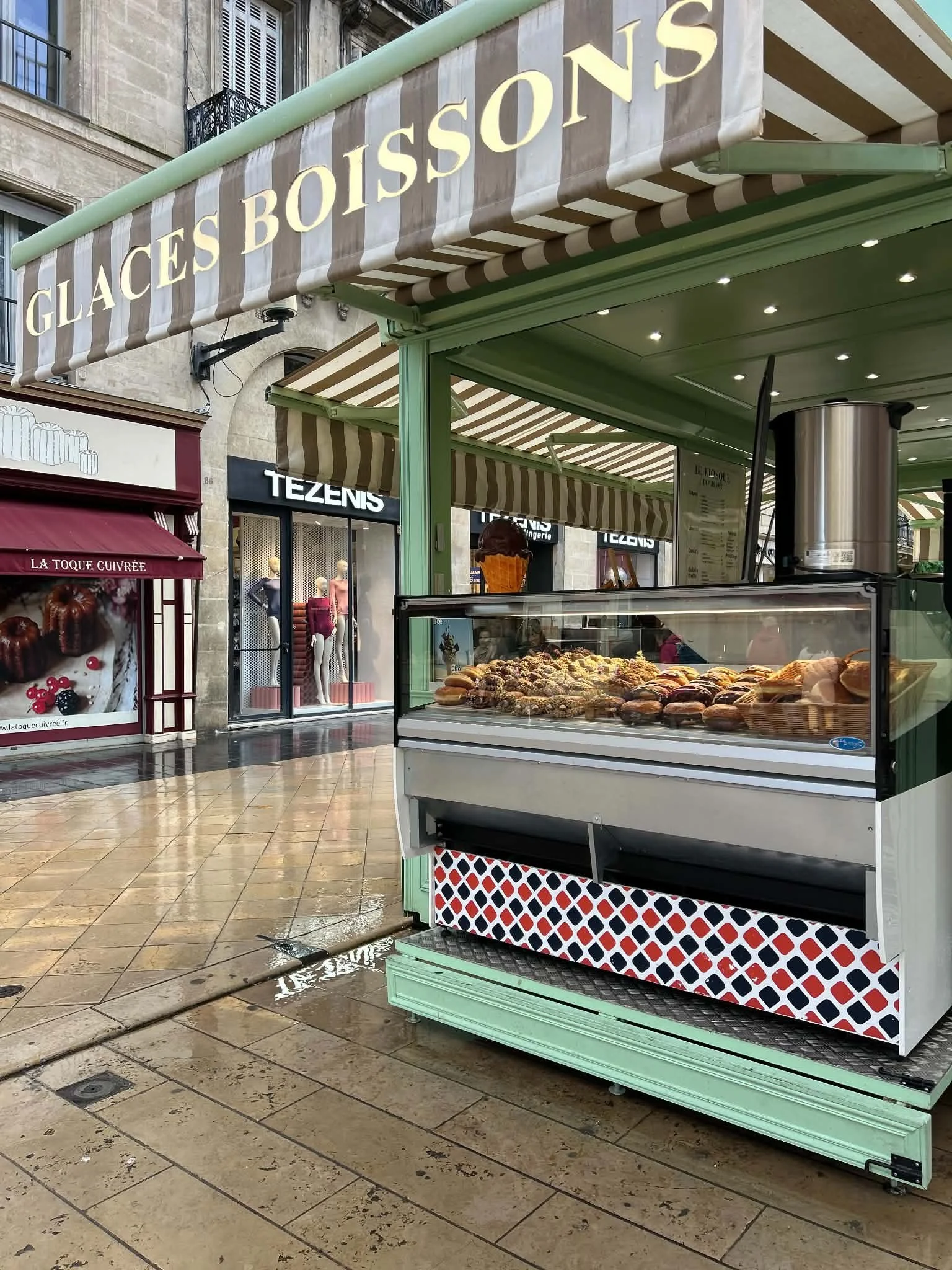 A street vendor cart with assorted baked goods, located in a shopping street with boutique stores and awnings, under cloudy weather with wet pavement.