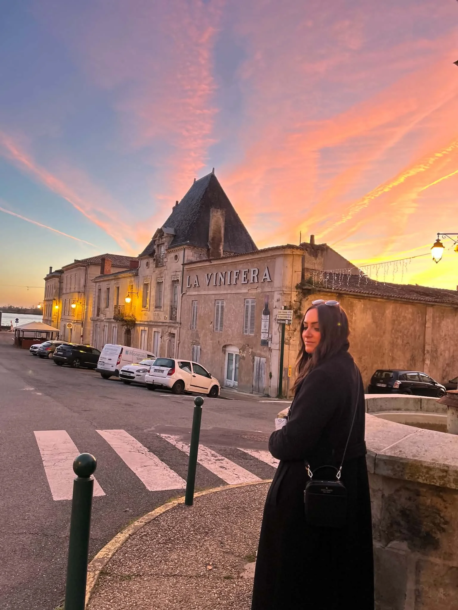 A woman standing on a street corner at sunset, with cars parked along the street and a historic building labeled 'La Vinfera' in the background, under a colorful sky with pink and orange hues.