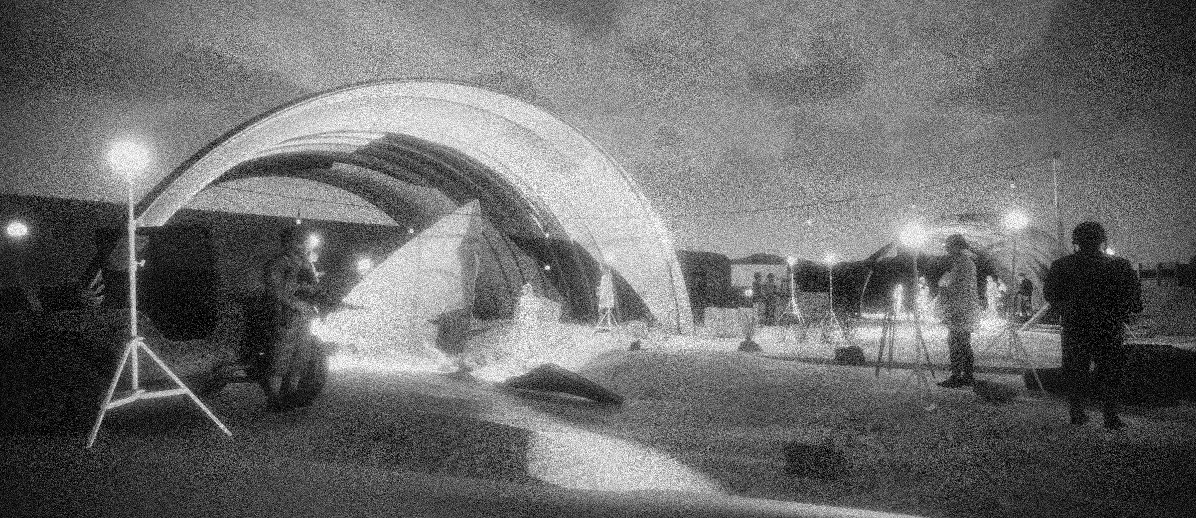 Night scene of a crew working with lighting and equipment under a large, illuminated, arched structure with several people present, some holding cameras or equipment, with a dark sky and distant horizon in the background.