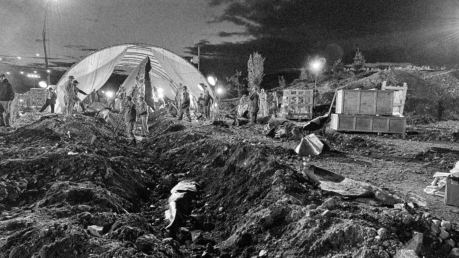 Nighttime scene of people working on a construction site with piles of dirt and rocks, some near a large tent structure, and equipment around, illuminated by floodlights.