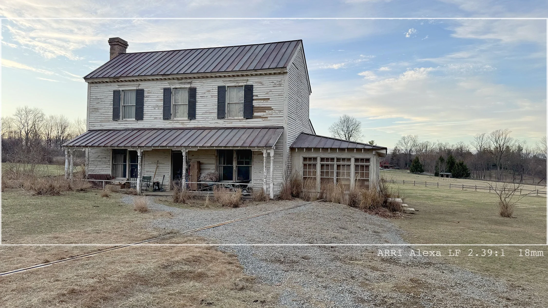 An old, weathered two-story house with peeling white paint and black shutters, sitting in a rural area with a gravel driveway, leafless trees, and open fields under a partly cloudy sky.