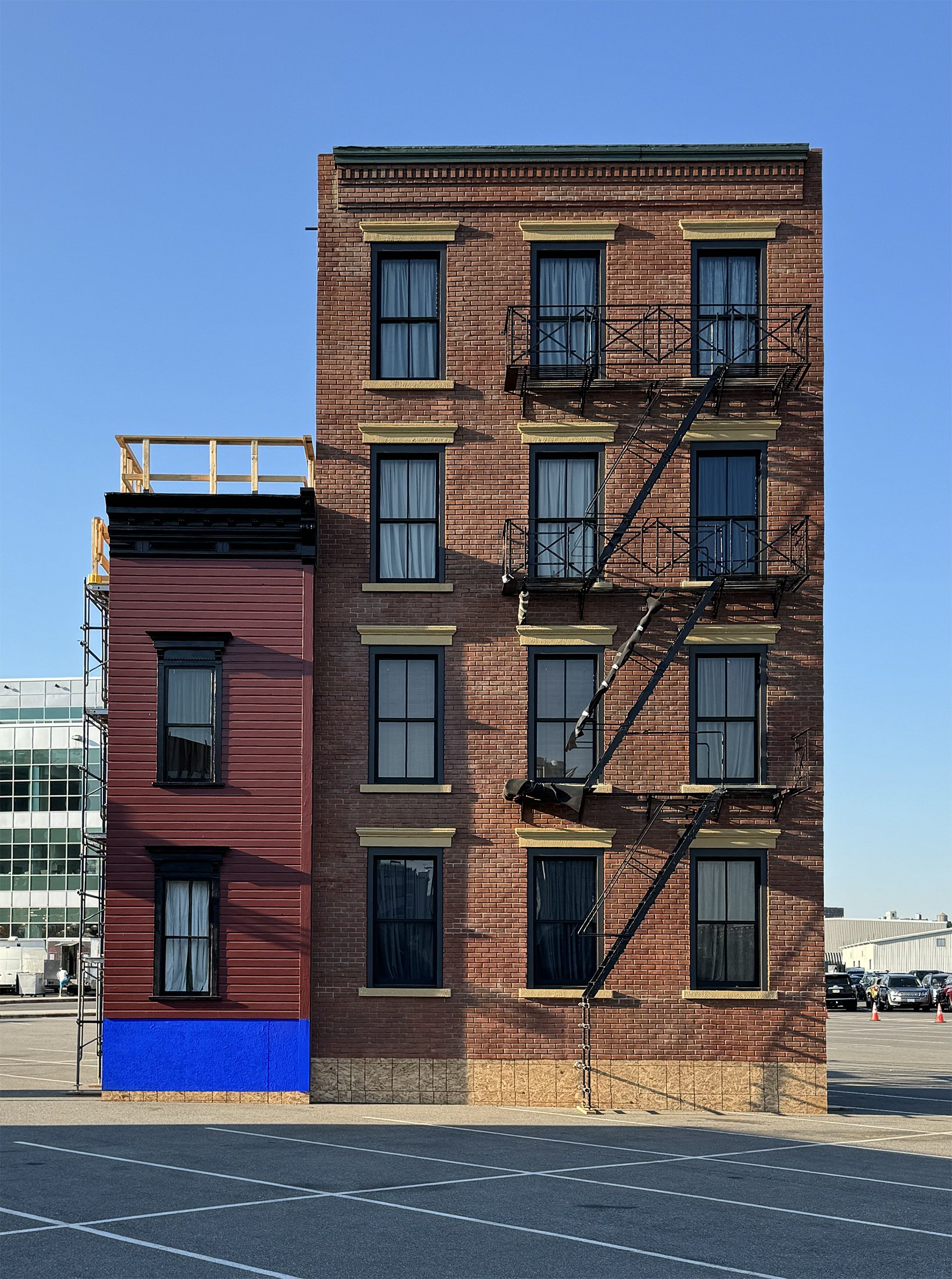 A multi-story brick building with fire escape stairs and balconies, partially painted blue on the lower left, against a clear blue sky, with a large parking lot in the foreground.