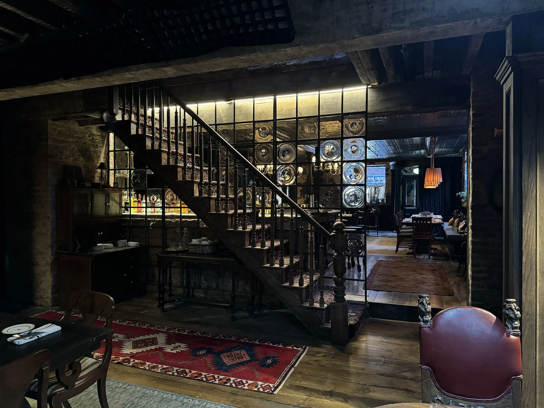 Interior of a cozy restaurant or cafe with wooden floors, brick walls, a staircase with a decorative railing, and a large grid window with glass bottles and decorative items inside. There's a patterned red and blue rug, a dining table with chairs and