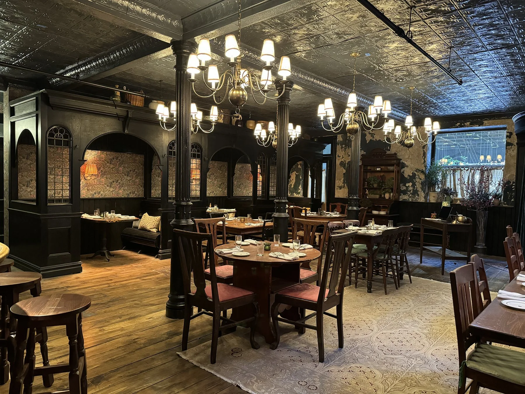 An elegant restaurant with dark wooden furniture, ornate chandeliers hanging from a decorative tin ceiling, and large windows letting in natural light. The space features a mix of round and rectangular tables set with white napkins, glassware, and pl