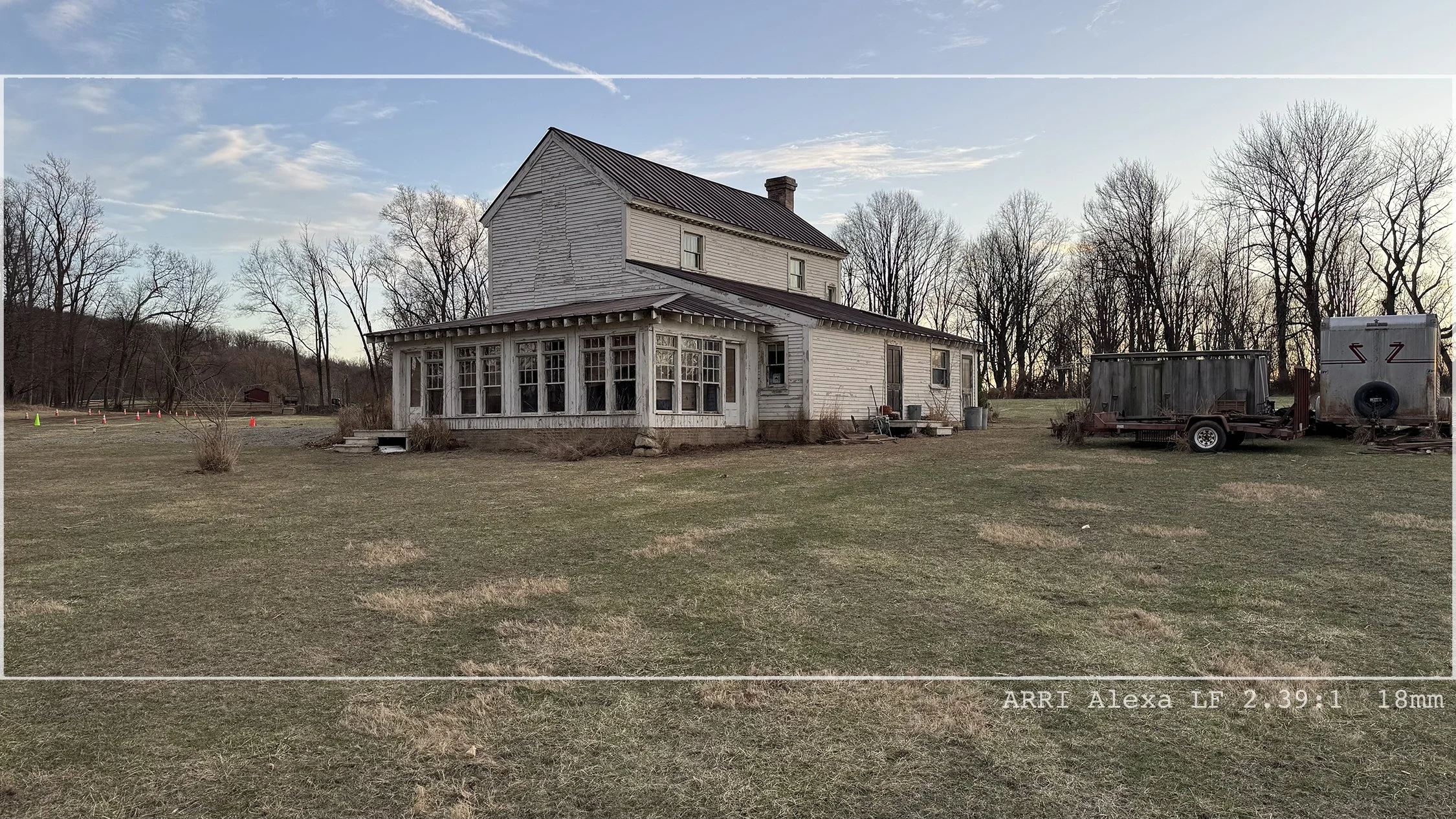 An old white wooden house with a screened porch on a grassy field, leafless trees in the background, and a trailer near the house. The sky is partly cloudy with faint contrails.