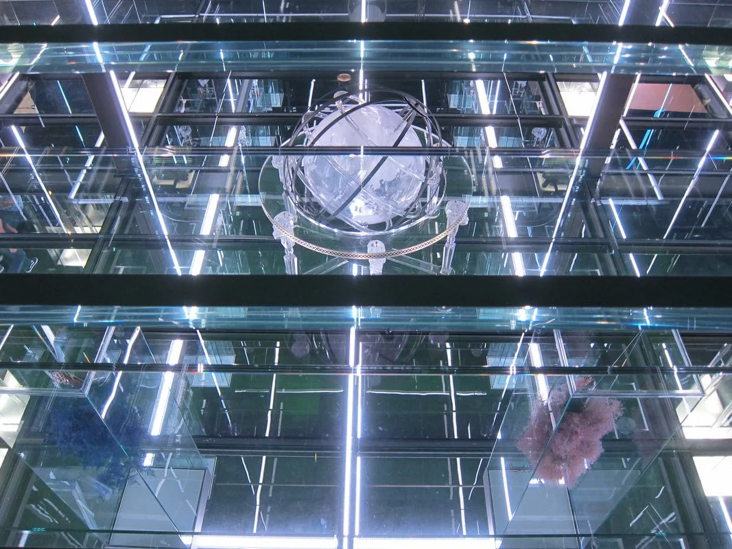 A modern building interior with multiple reflections, illuminated by bright white LED lights, featuring a glass and metal globe sculpture hanging in the center.