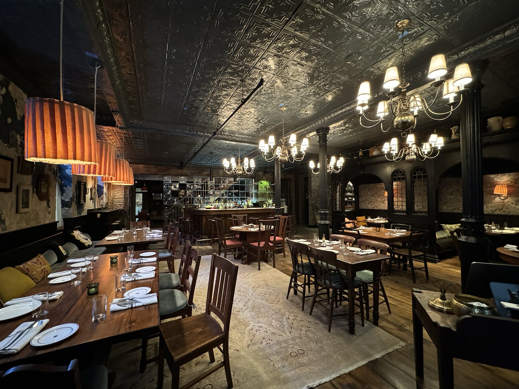 Interior of a restaurant with wooden tables set with white plates, glasses, and napkins. The ceiling is decorative with metallic panels, and the room is illuminated by multiple chandeliers and hanging lamps. There are couches with cushions along one 