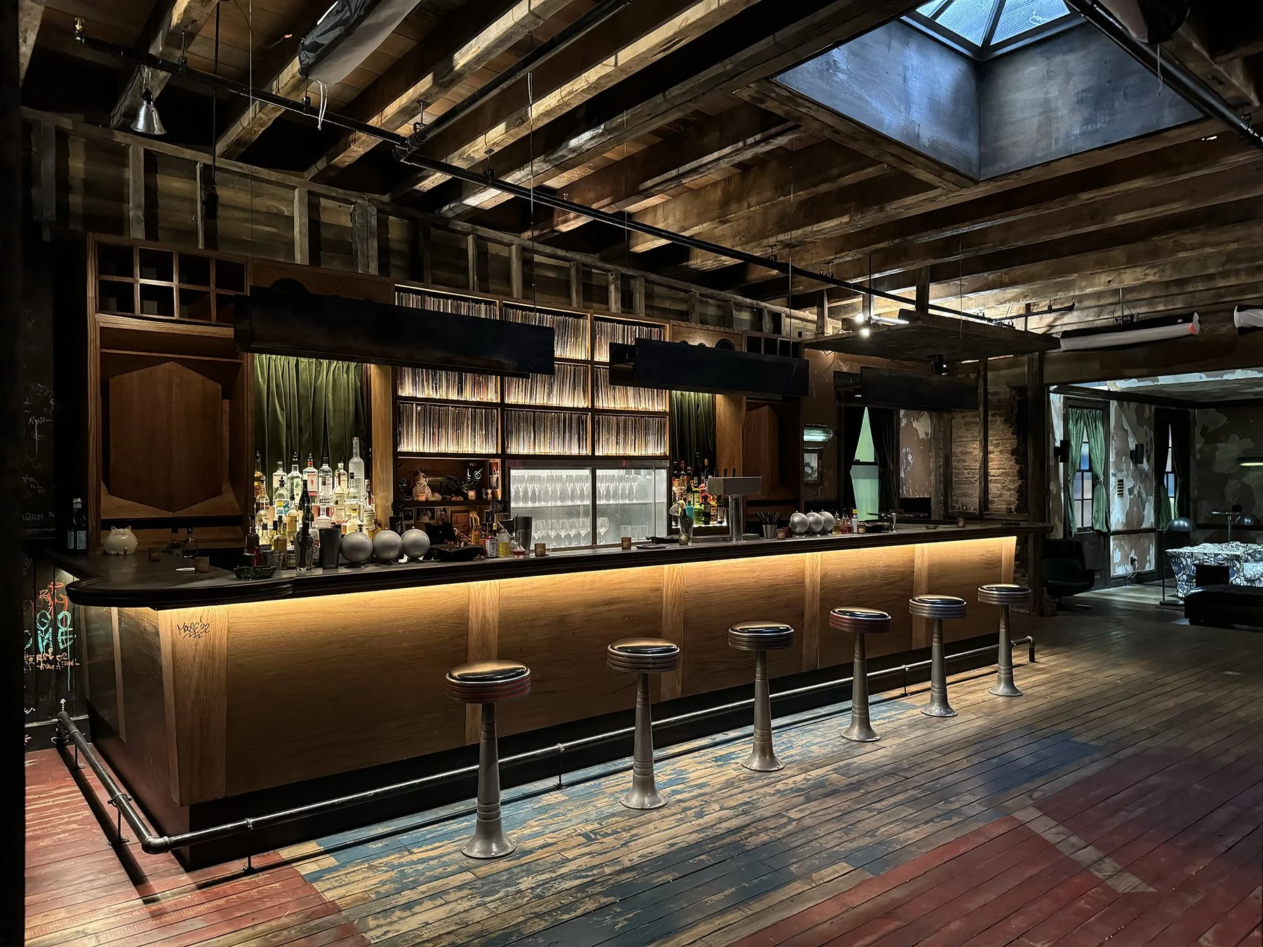 Empty bar with a wooden counter, liquor bottles and glasses, surrounded by barstools, in a rustic, dimly lit, industrial-style interior with exposed wood beams.