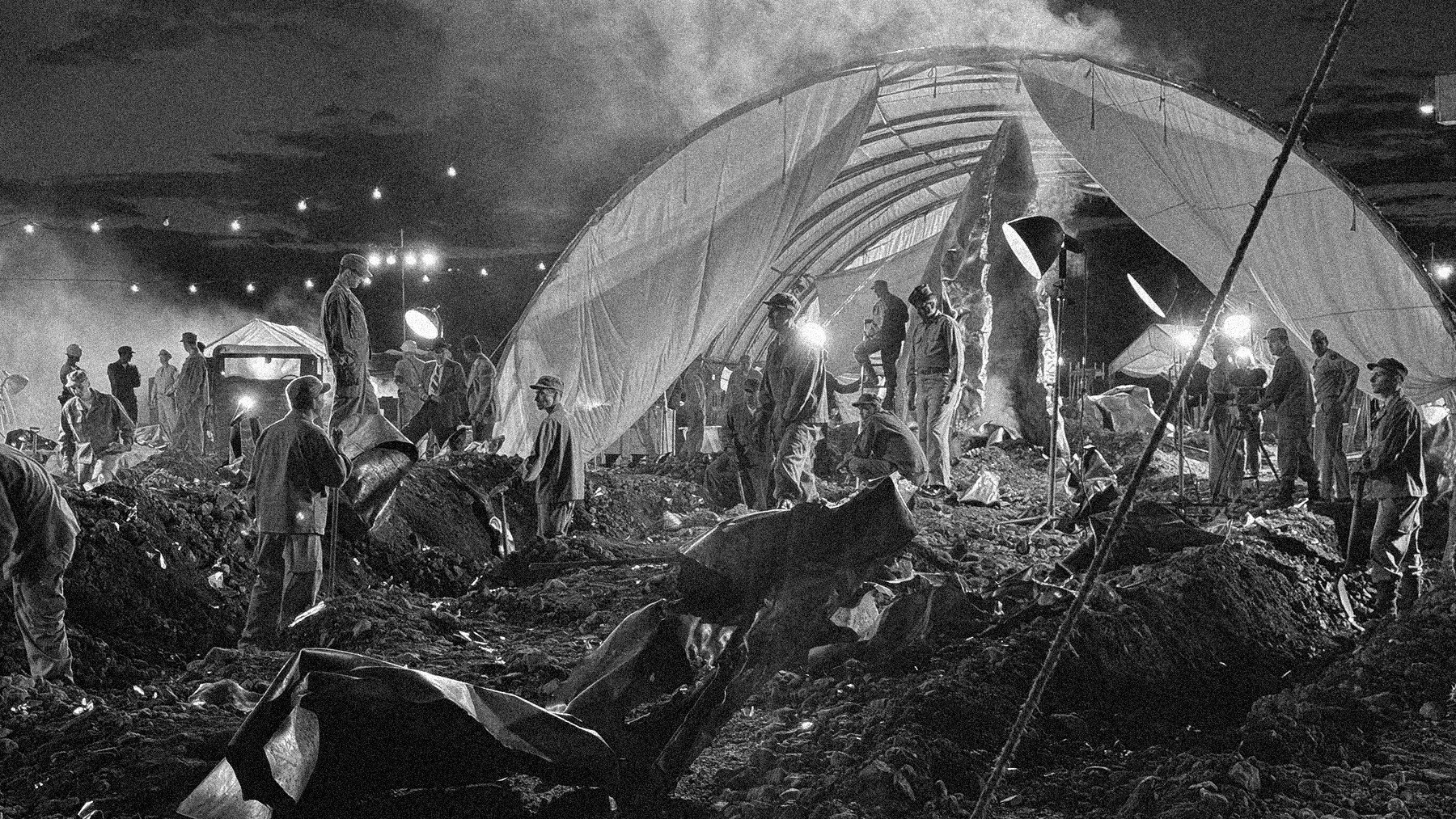 Black and white photograph of an excavation site at night with workers moving dirt and debris under large tent-like structures, illuminated by bright lights.