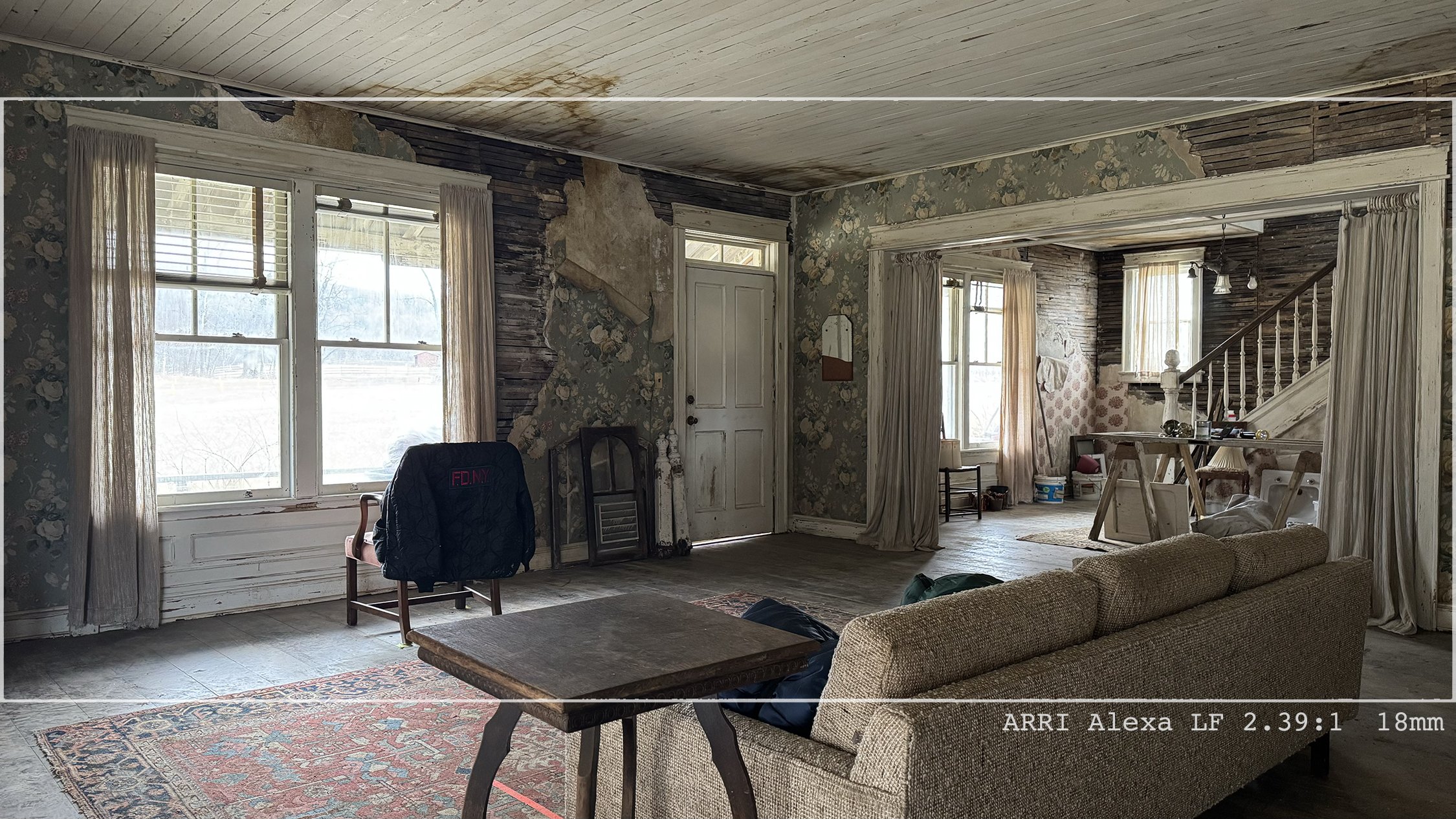 Interior of an old, worn house with peeling wallpaper, damaged walls, and weathered wood ceiling. Large windows with curtains, a couch, a coffee table, and various furniture and belongings scattered around.