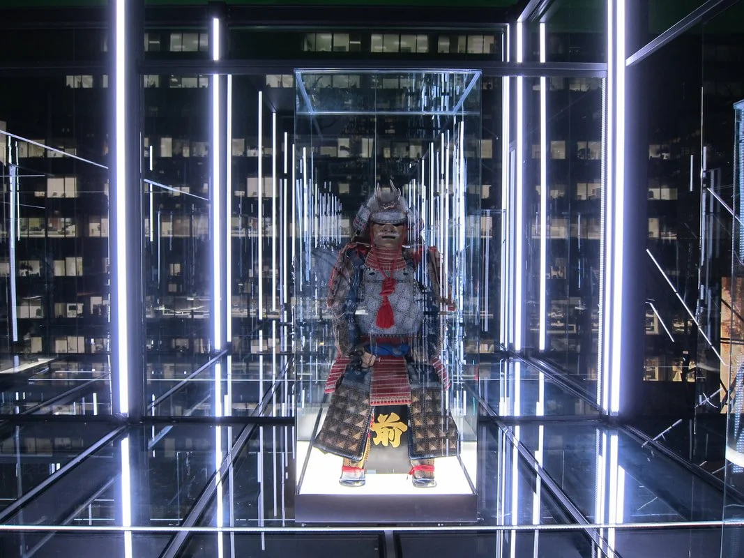 A glass display case with a traditional Japanese samurai armor set inside, illuminated by bright white LED lights, situated in an modern architectural setting.