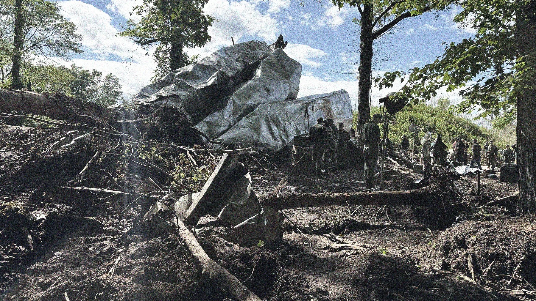 Scene of destruction with large wreckage covered in a tarp, debris, fallen trees, and a group of people and rescue workers amidst the aftermath of a disaster in a forested area.