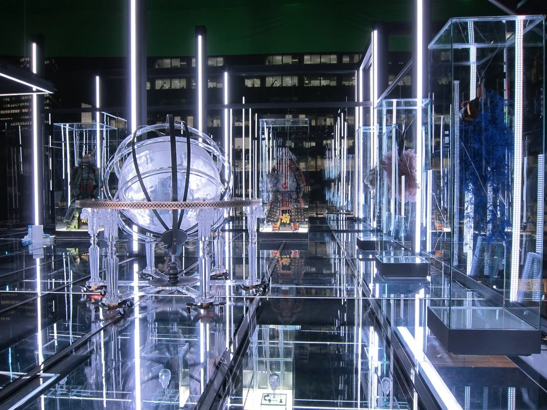 Futuristic display with illuminated glass cases showcasing mannequins and objects, featuring bright white and blue neon lights, on a reflective floor in a modern exhibition or store.