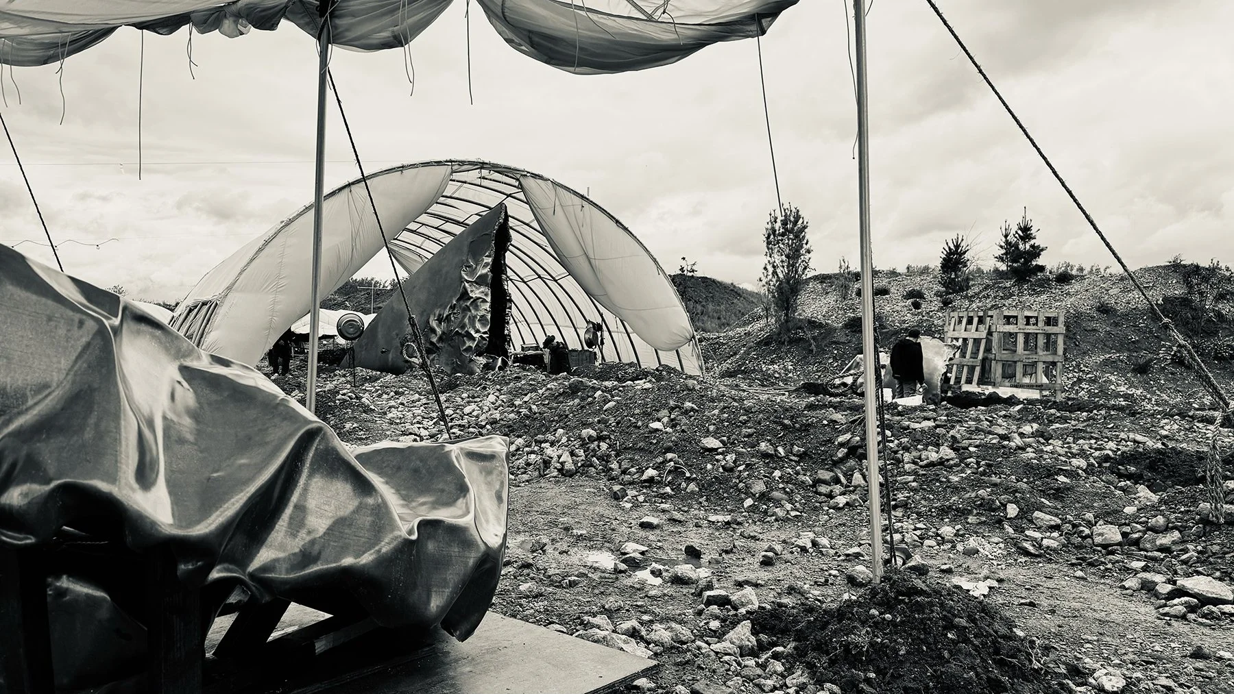 A photo of a partially destroyed greenhouse with a damaged structure, surrounded by rocky terrain. There are some small trees in the background, and a couple of people are visible walking among the debris and construction materials.