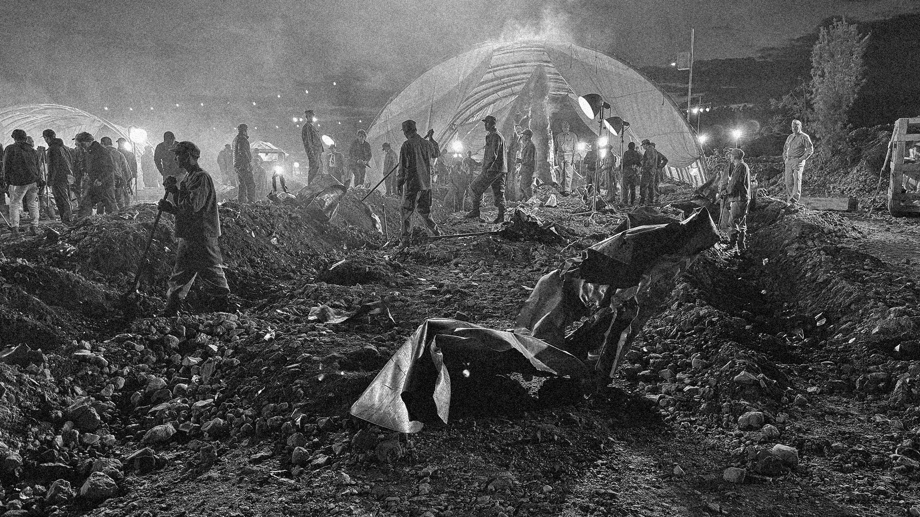 Nighttime scene of a construction or excavation site with many workers, some shoveling dirt and rocks. Bright lights illuminate the area, and a large tent or structure is visible in the background. The ground is uneven and filled with rocks, and vari