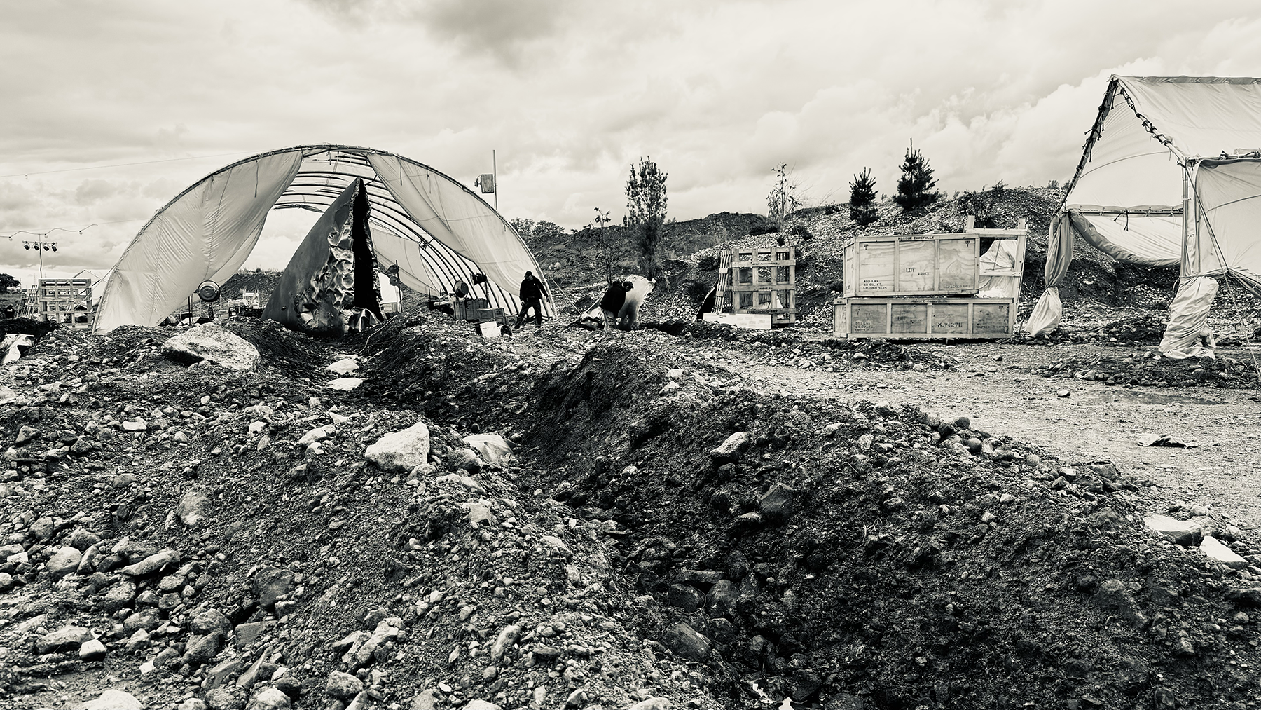 A black and white photo of a barren outdoor scene with large rocks and dirt mounds. There are tents or temporary structures, some damaged, with a few people working or walking around. Wooden crates and construction materials are scattered, with overc