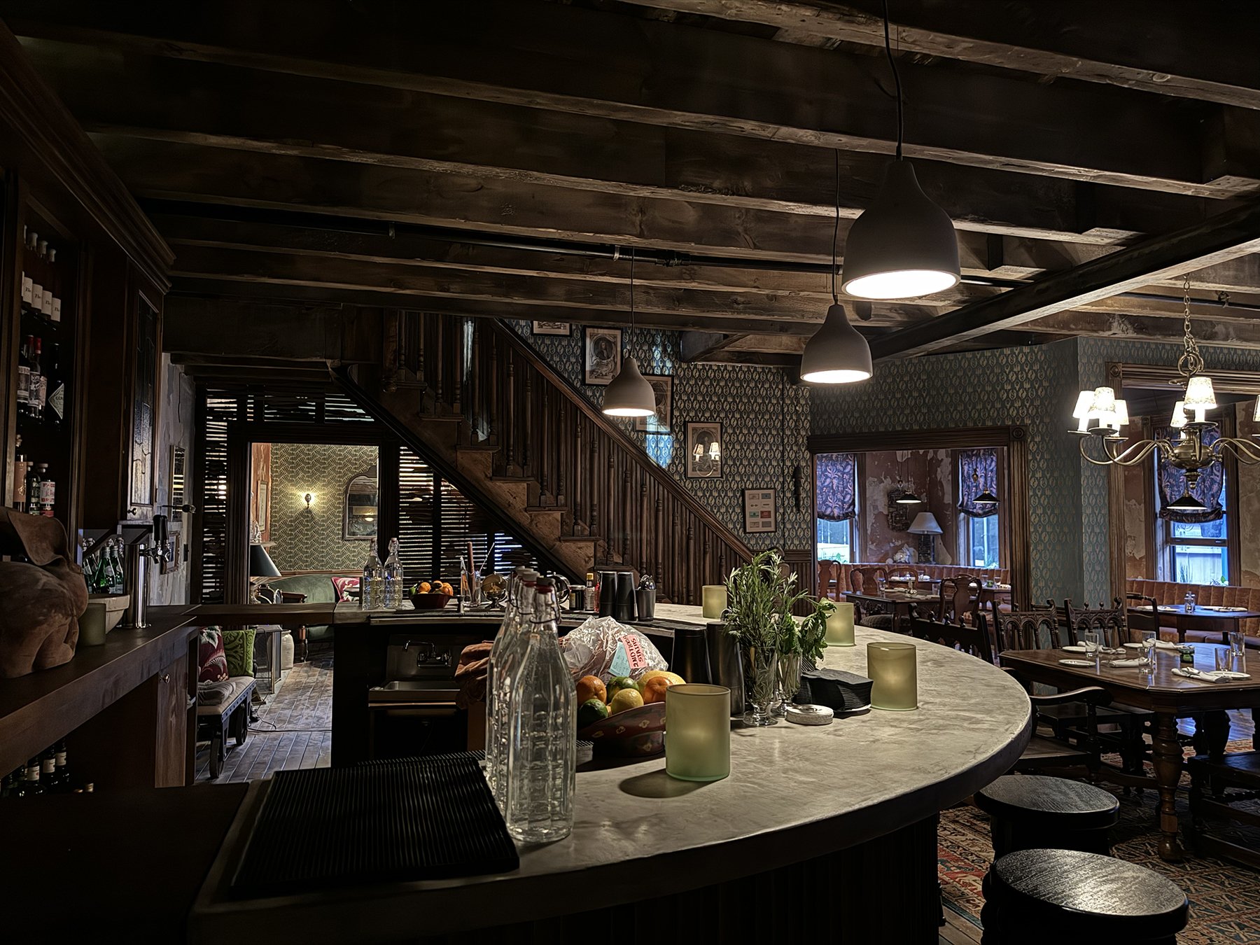 Interior of a cozy restaurant with a marble counter, wooden staircase, and vintage decor, including patterned wallpaper, hanging pendant lights, and a chandelier, with tables set for dining.