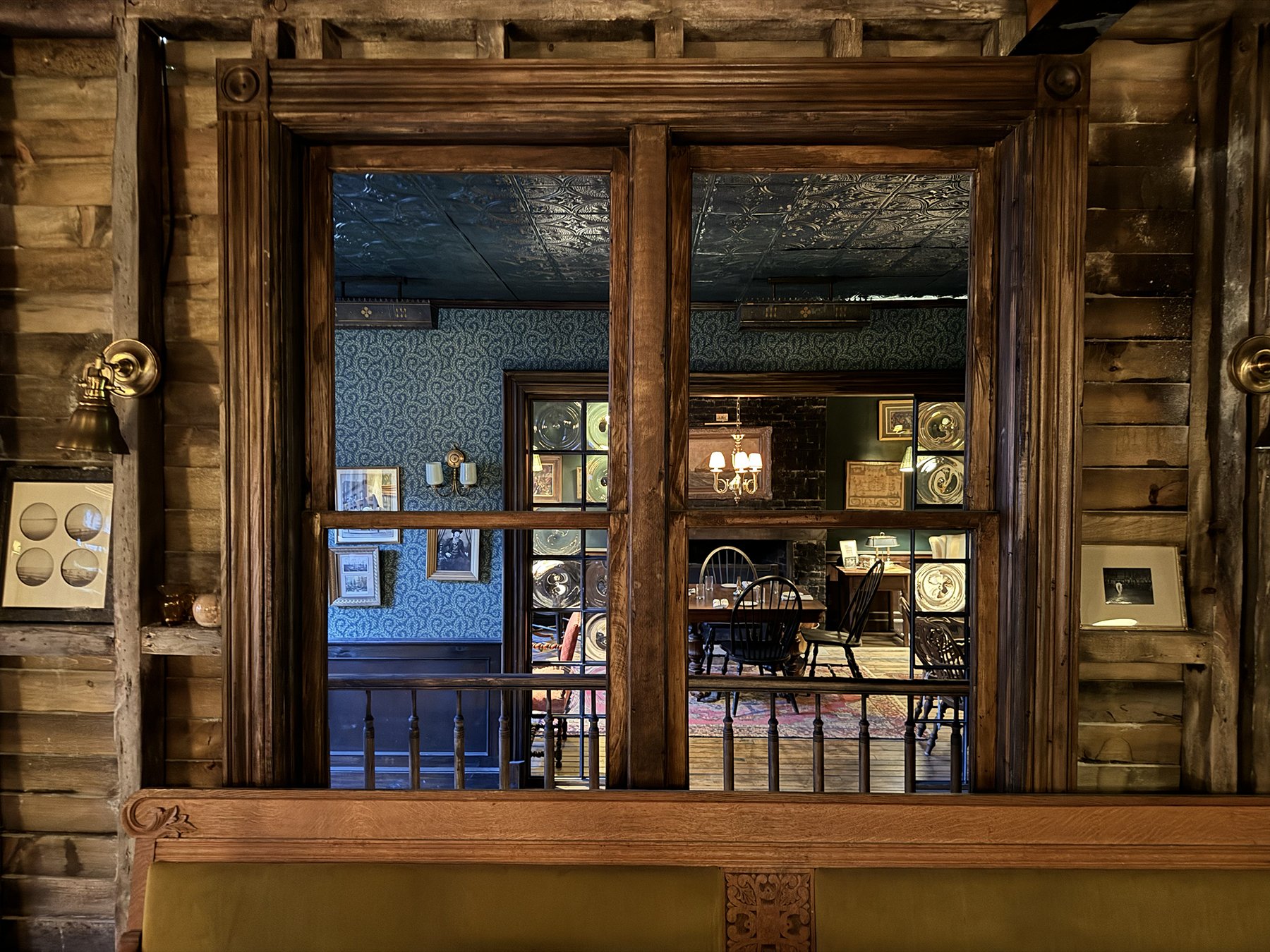 View through a wooden framed window into a cozy living room with dark patterned wallpaper, framed art, a chandelier, and a dining table with chairs.