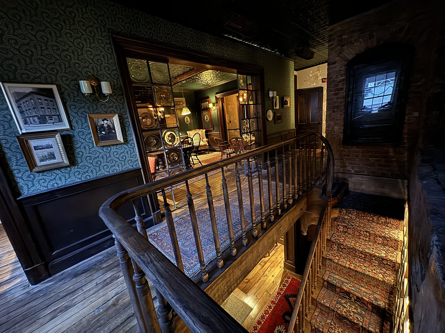 Interior view of a vintage house with wood and brick walls, patterned carpets, framed photographs, and decorative plates, with a hallway leading to a room with a dining area and more vintage decor.
