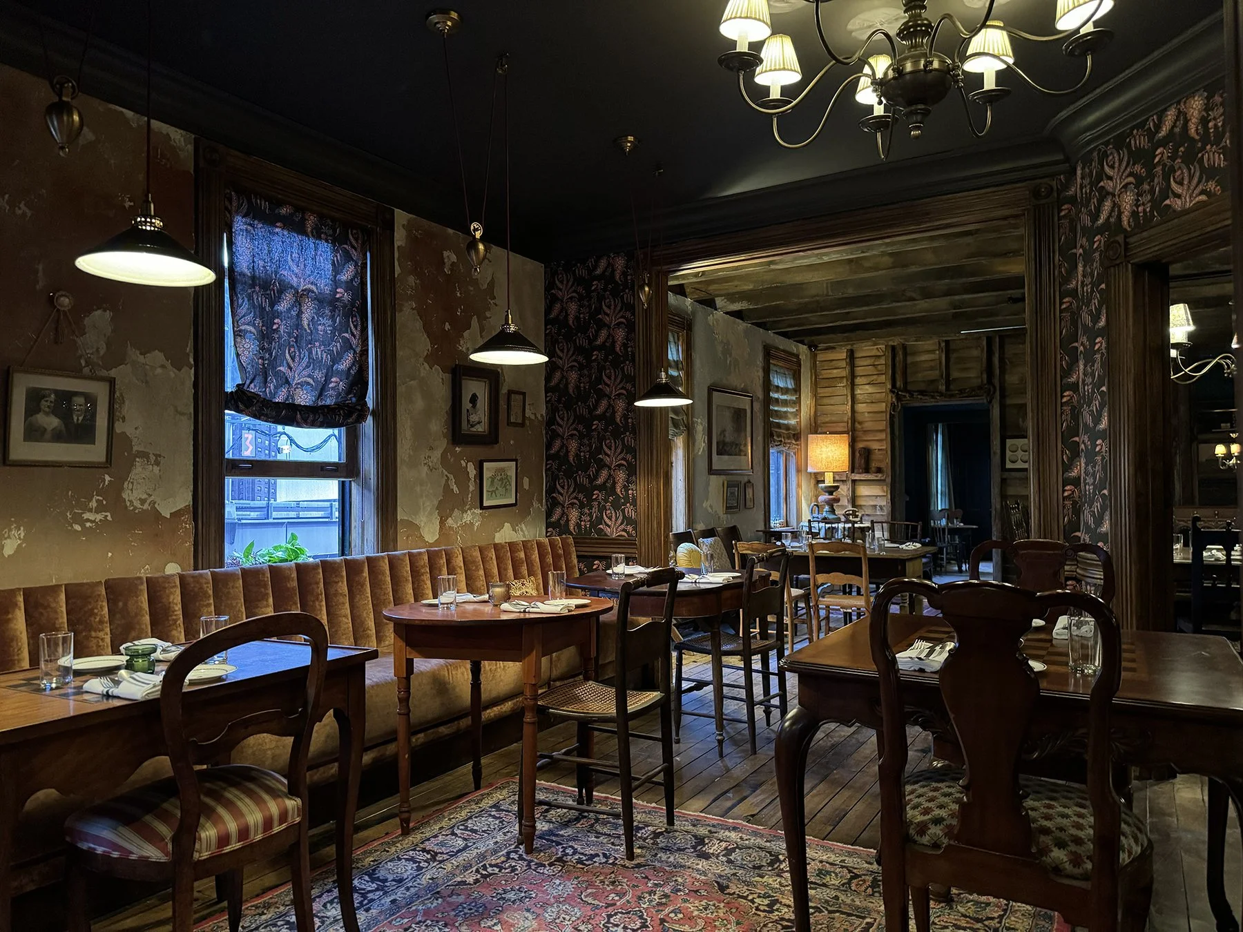 Interior of a cozy vintage-style restaurant with wooden furniture, pendant and chandelier lighting, patterned wallpaper, and framed artwork on weathered walls. Dining tables are set with glasses, plates, and napkins, and the space features large wind