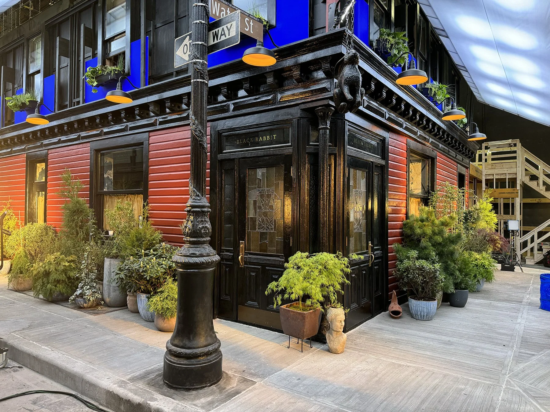 Street corner with red and black building, surrounded by potted plants, street signs, and a black lamppost.