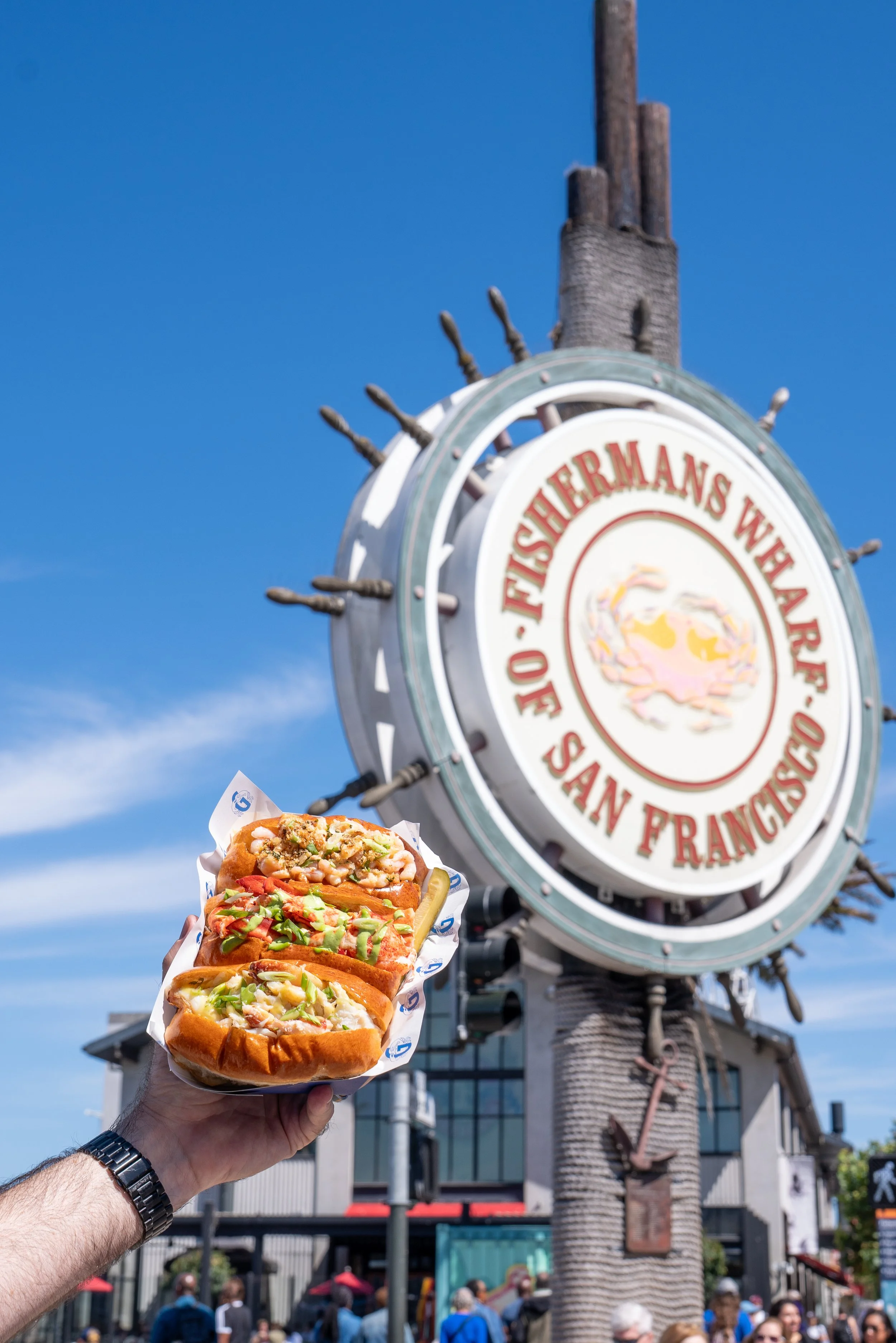 Trio of crab, lobster, and shrimp rolls held up in front of the Fisherman’s Wharf sign in San Francisco
