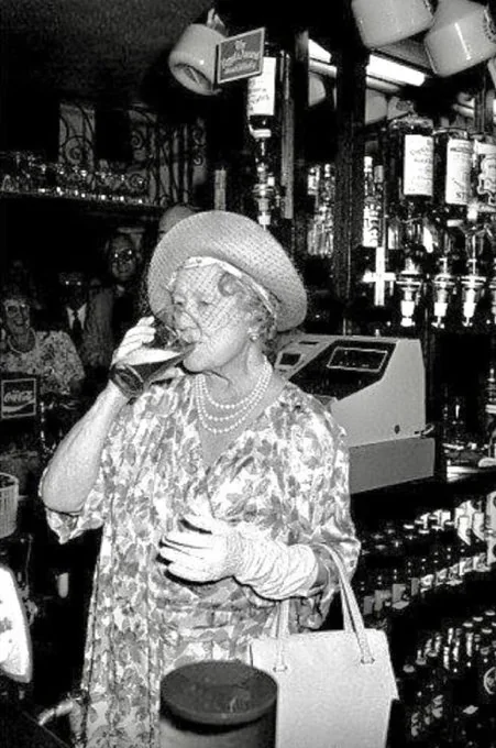 The Queen's Mother, An elderly woman in vintage attire, including a hat, pearl necklaces, and gloves, drinking a pint of bitter, behind the bar at the Queen's Head.