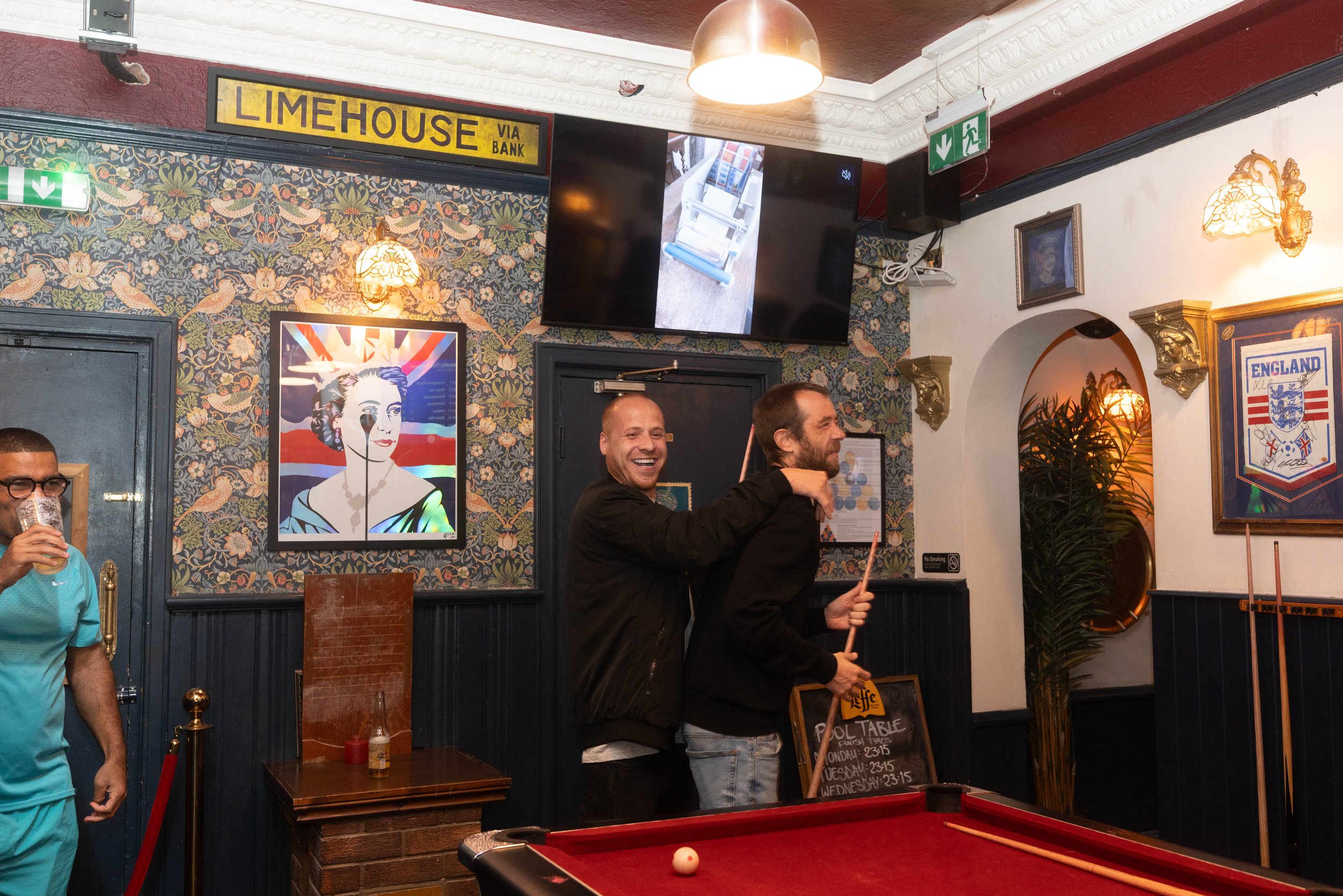 People playing pool in a bar or pub, with a pool table in the foreground and a television on the wall and framed pictures in the background.