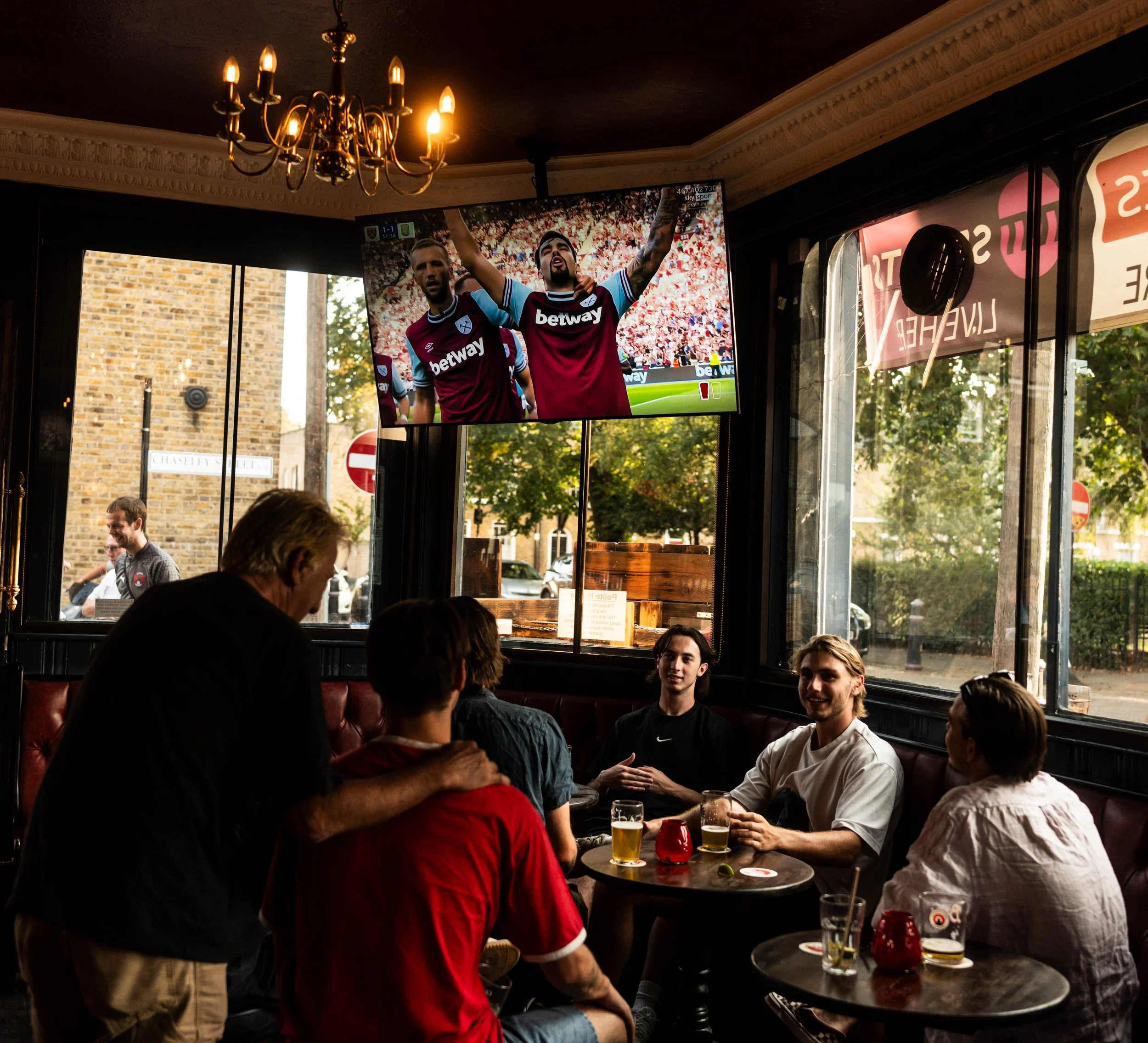 People watching a football game on a large TV inside a restaurant, with some holding drinks and chatting, in a cozy interior with chandelier and large windows showing an outdoor scene.