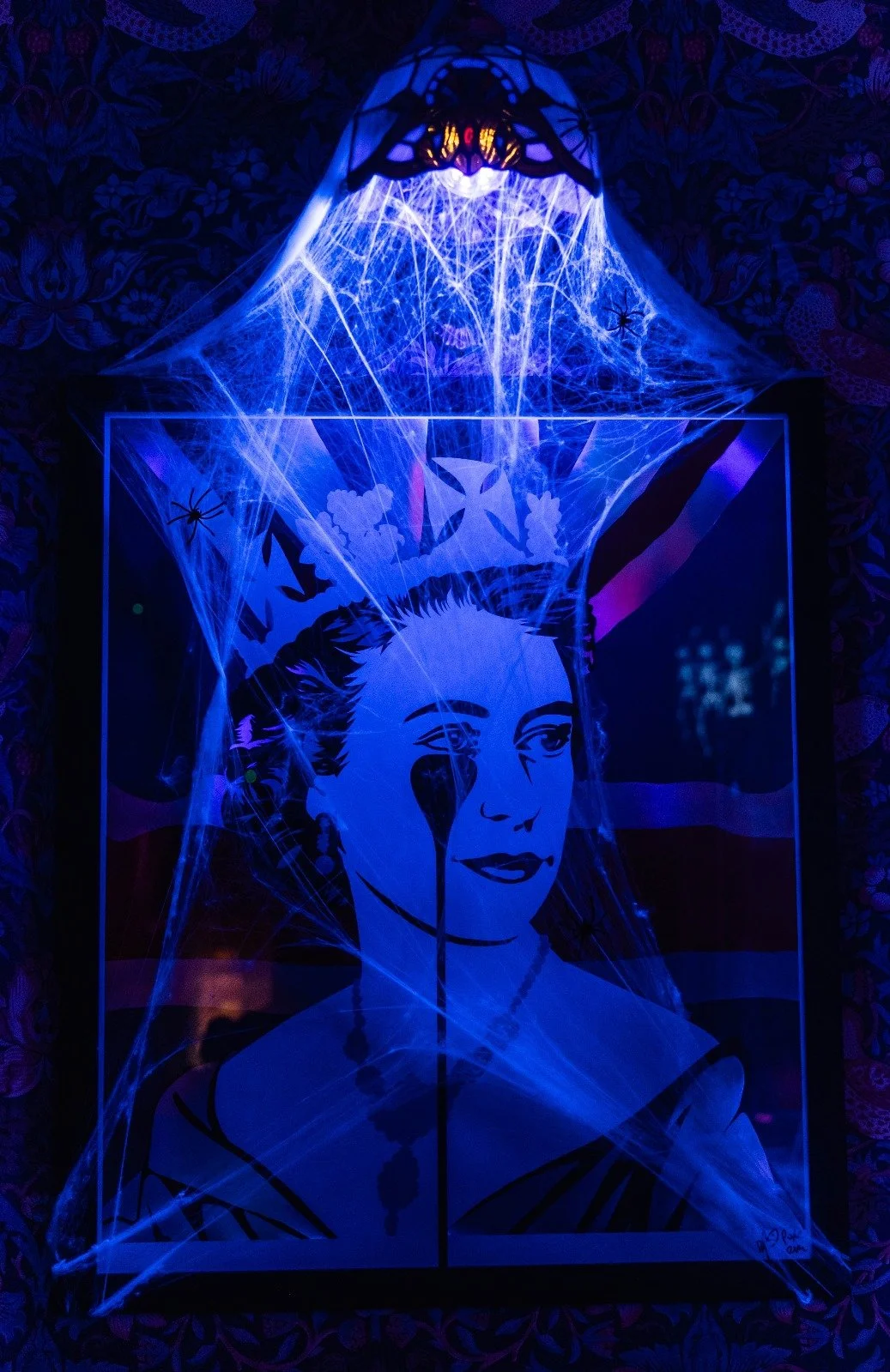 Illuminated artwork of a woman with an intense gaze, surrounded by spider webs and spiders, with a background featuring the American flag and a crown on her head under a dark, patterned ceiling.