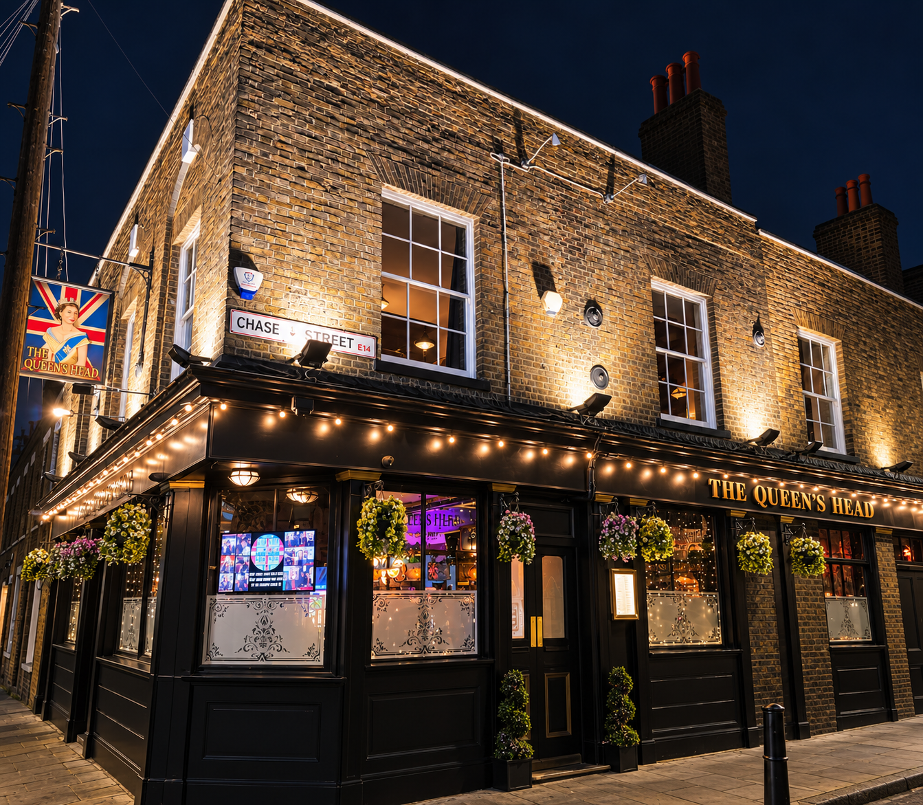 Exterior of a pub called The Queen's Head at night with string lights, hanging flower baskets, and illuminated windows on a brick building located on Chaseley Street.