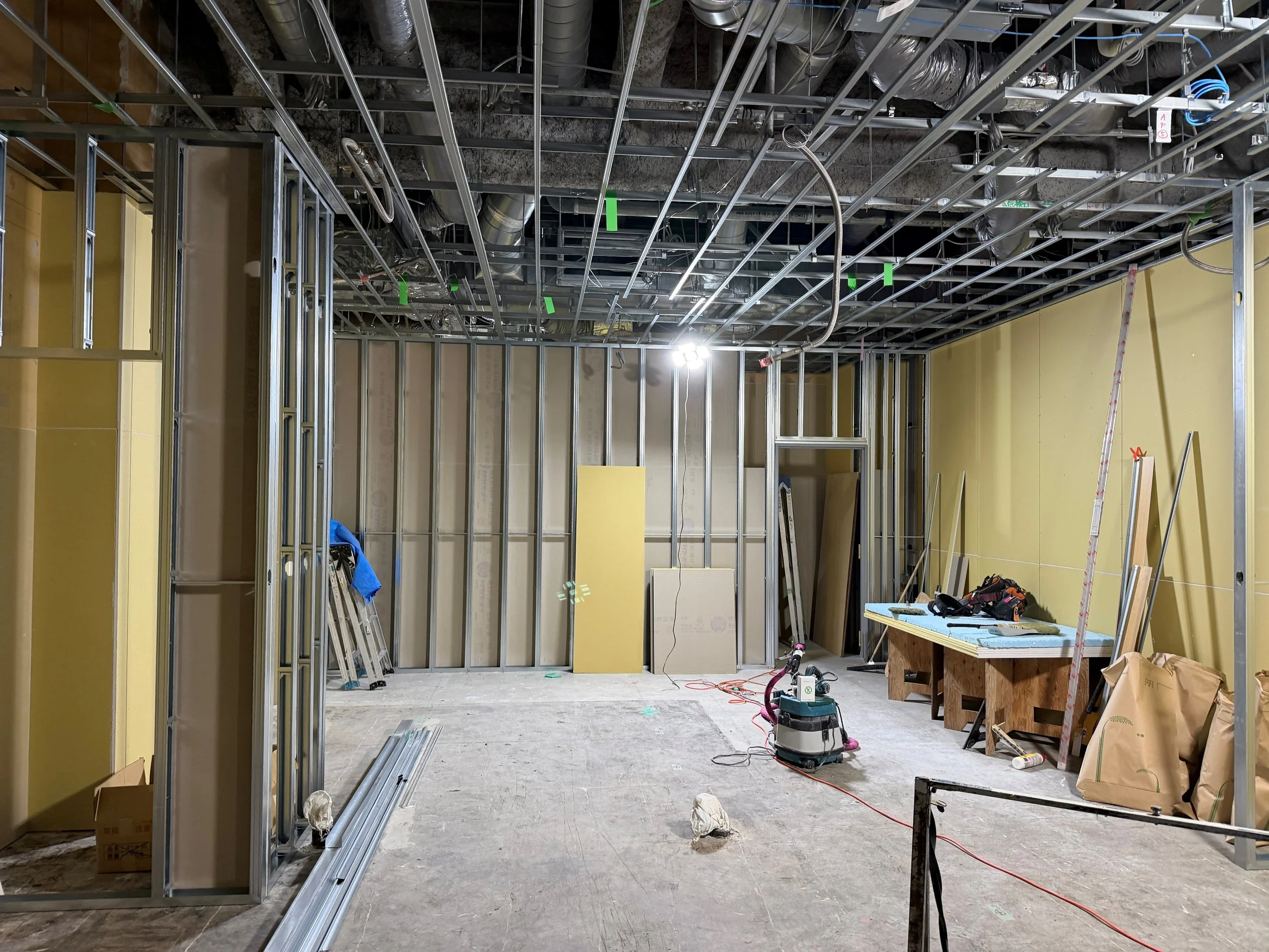 内装工事中の建物内部。軽量鉄骨の間仕切り壁、石膏ボード、露出した空調ダクトや配線、脚立と工具が見える。
