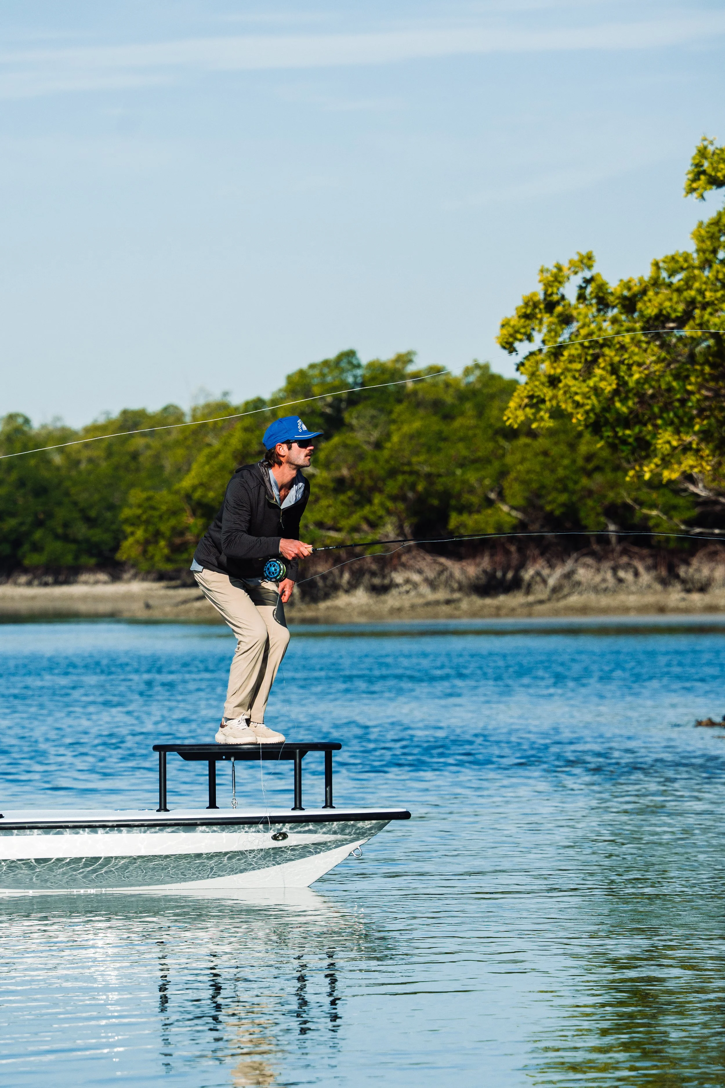 A man standing on a small platform on a boat, fishing in a body of water with green trees in the background.