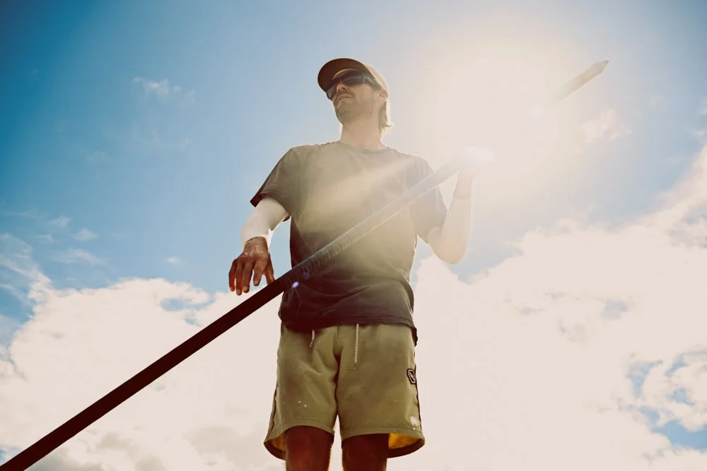 A man wearing a cap and sunglasses holding a paddle, standing outdoors in bright sunlight against a cloudy sky.
