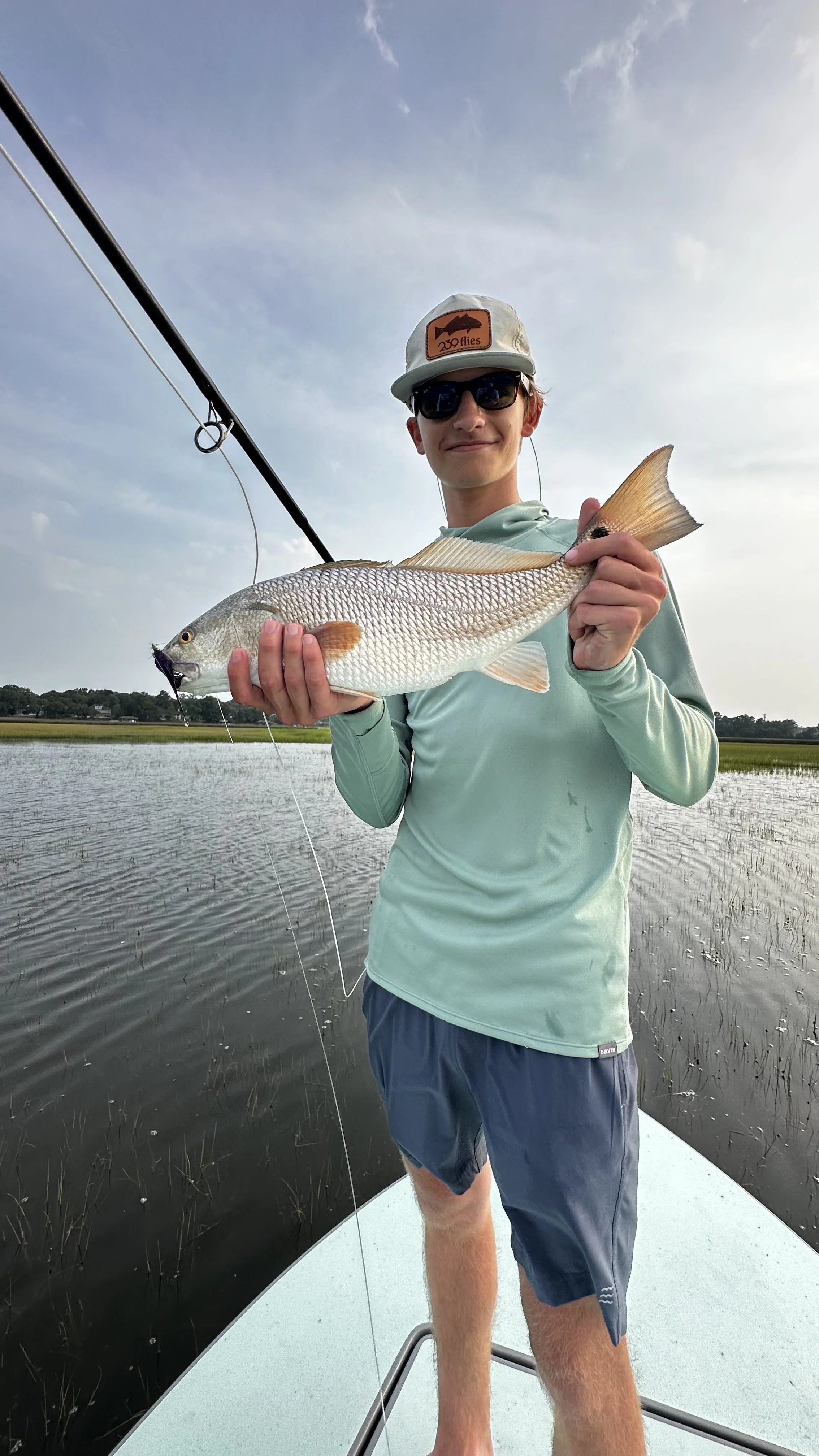 Young man wearing sunglasses, a cap with an orange patch, and fishing in a body of water, holding a large fish.