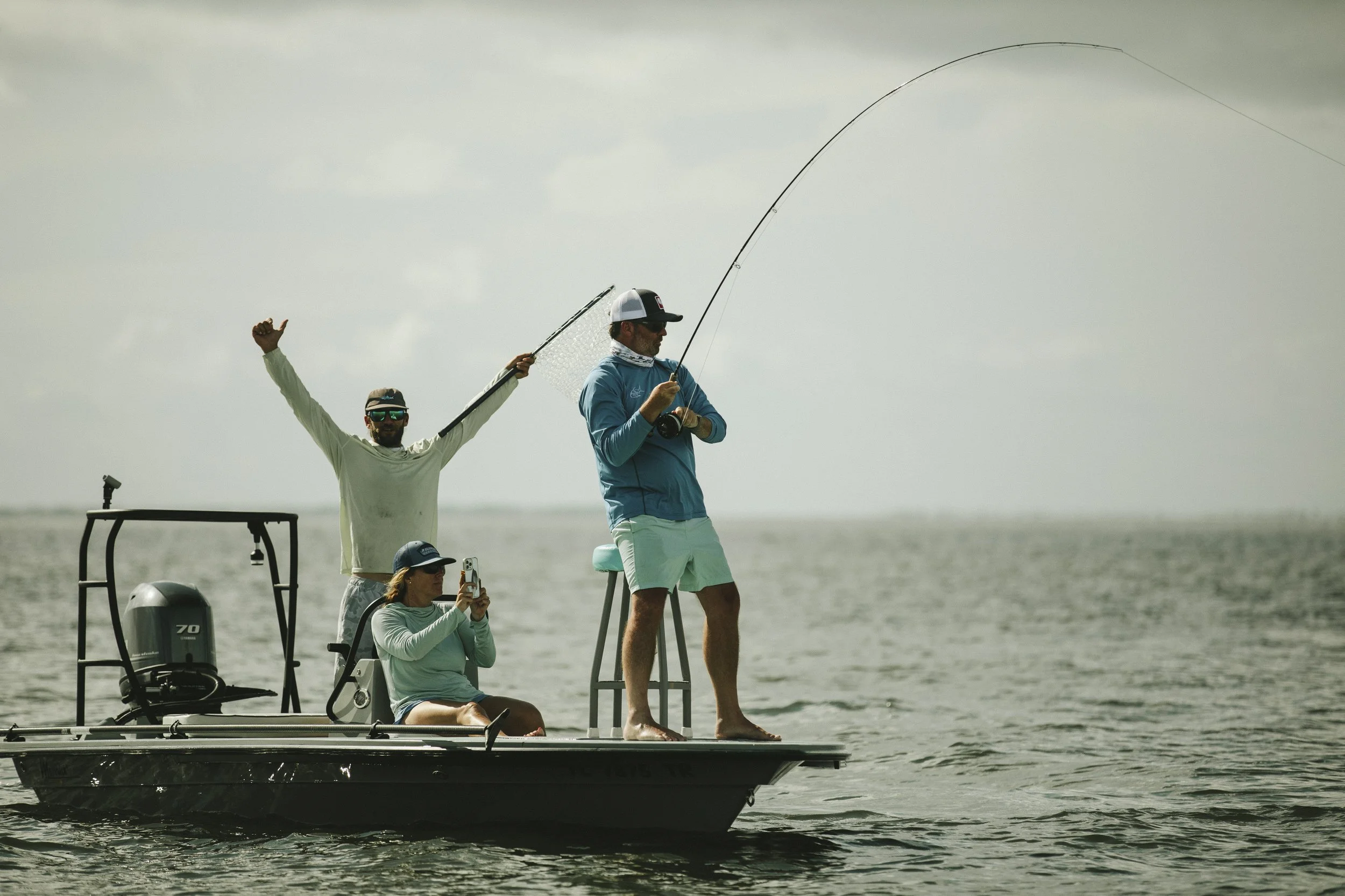A group of people fishing from a boat on a lake, with one person casting a fishing line and others watching or recording.