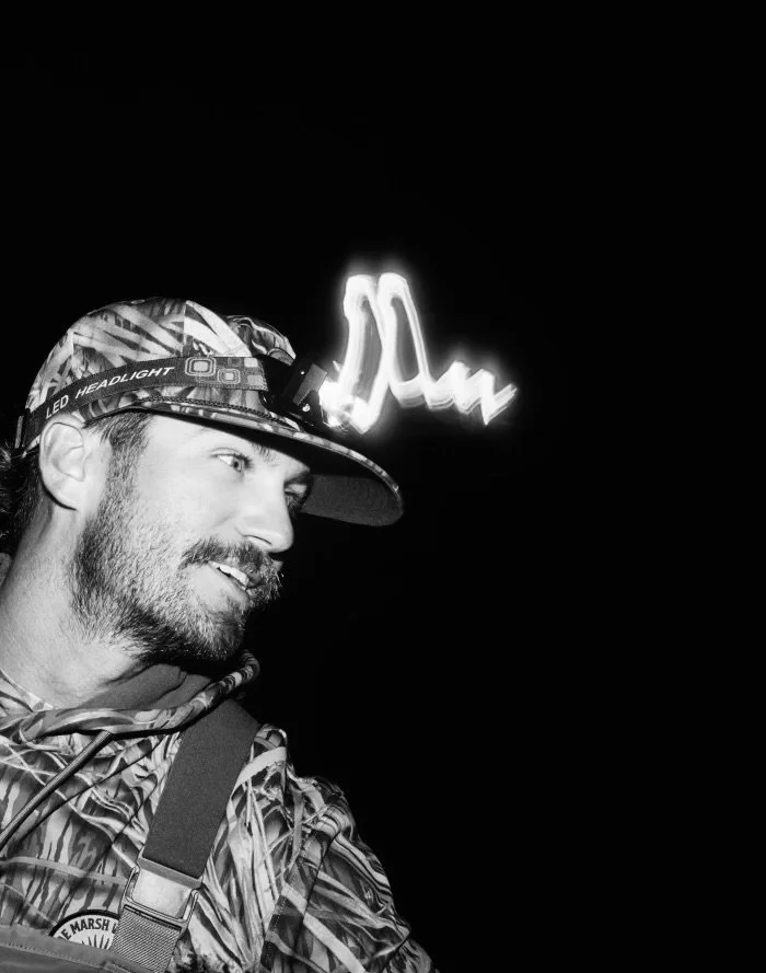 A man wearing a camouflage hat with an LED headlamp, a patterned jacket, and a backpack, smiling and looking to the left against a dark background with a neon sign.