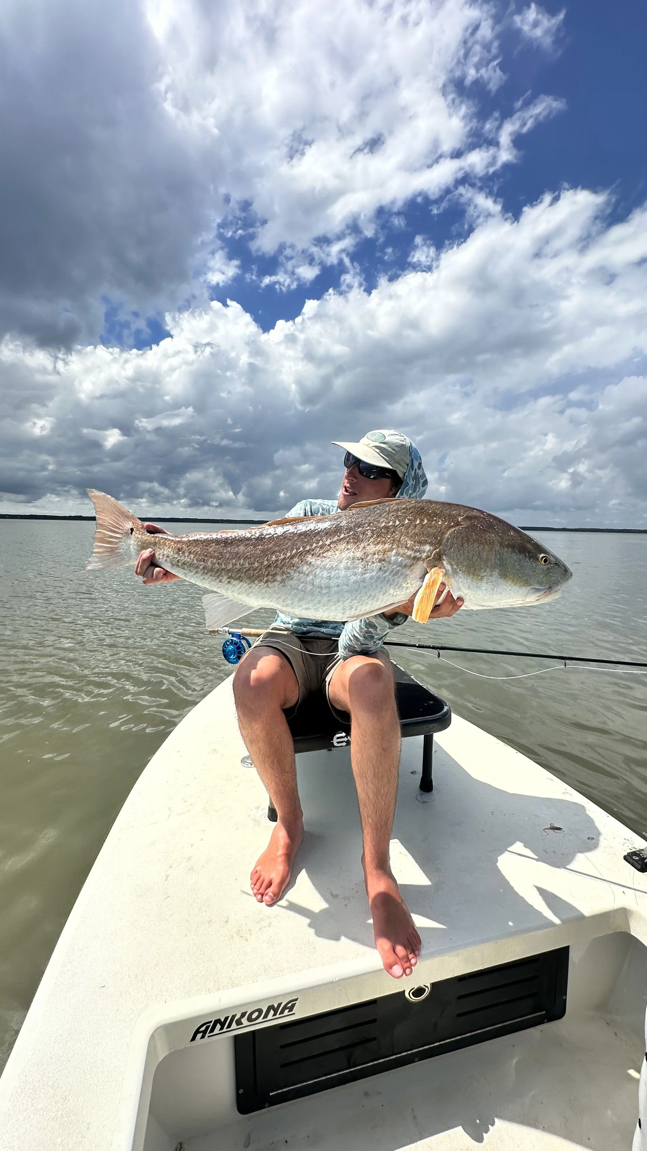 Person sitting on a boat holding a large fish in both hands. The person is wearing sunglasses, a hat, and casual clothing. The background shows water and a partly cloudy sky.