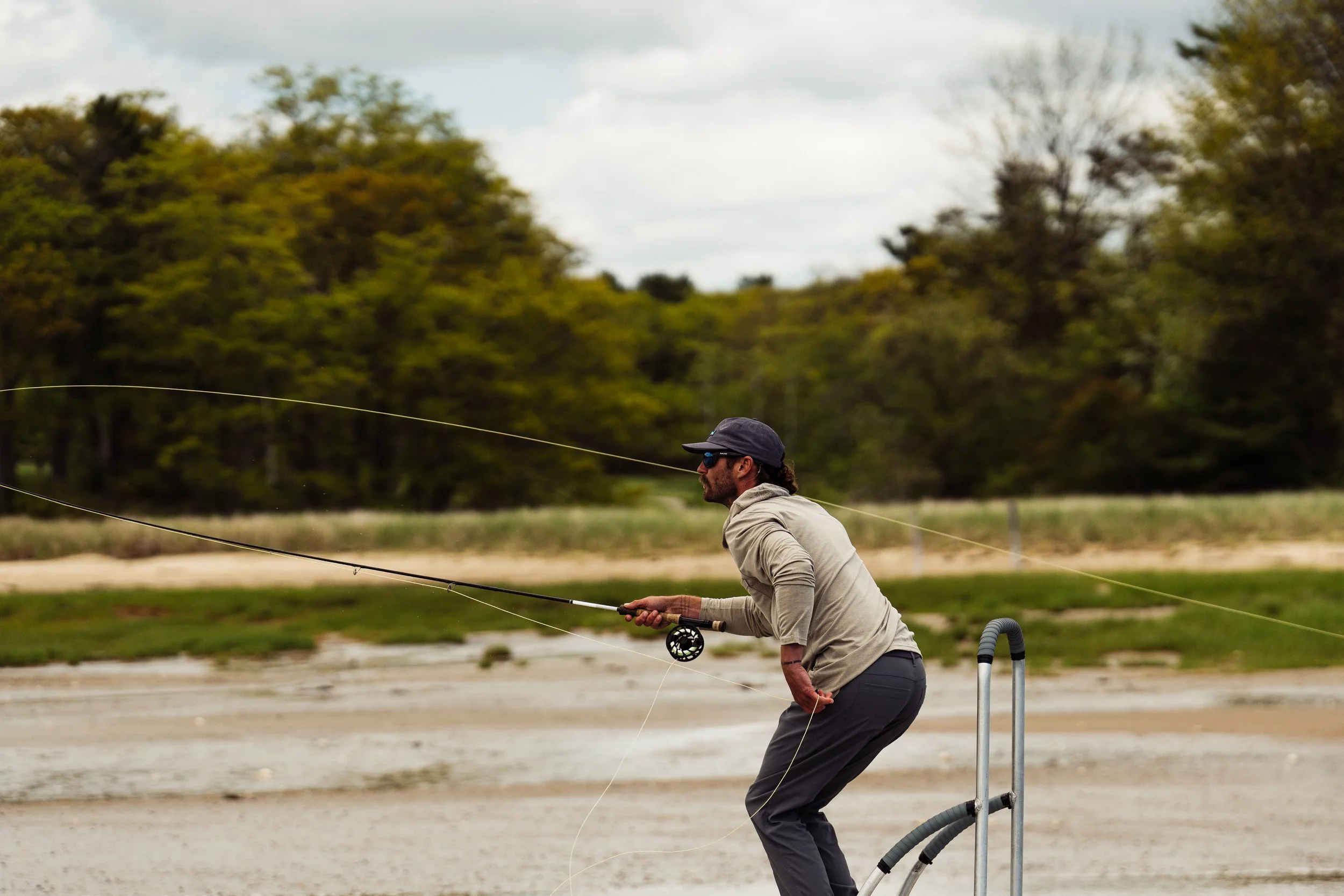 A man wearing sunglasses, a gray hat, a gray long-sleeve shirt, and gray pants is fishing in a river or lake, casting his fishing line. The background shows trees and a cloudy sky.