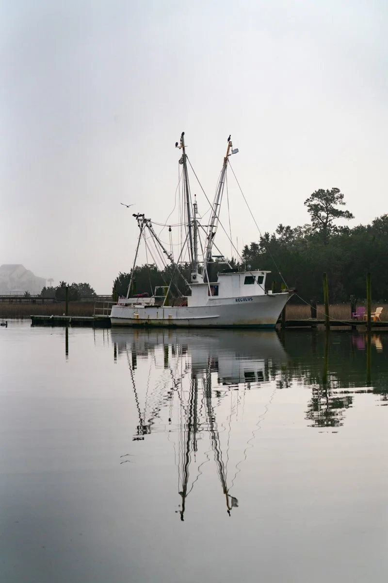 A white fishing boat named 'Regulus' docked on calm water, with its reflection visible below. The background shows trees and overcast sky.