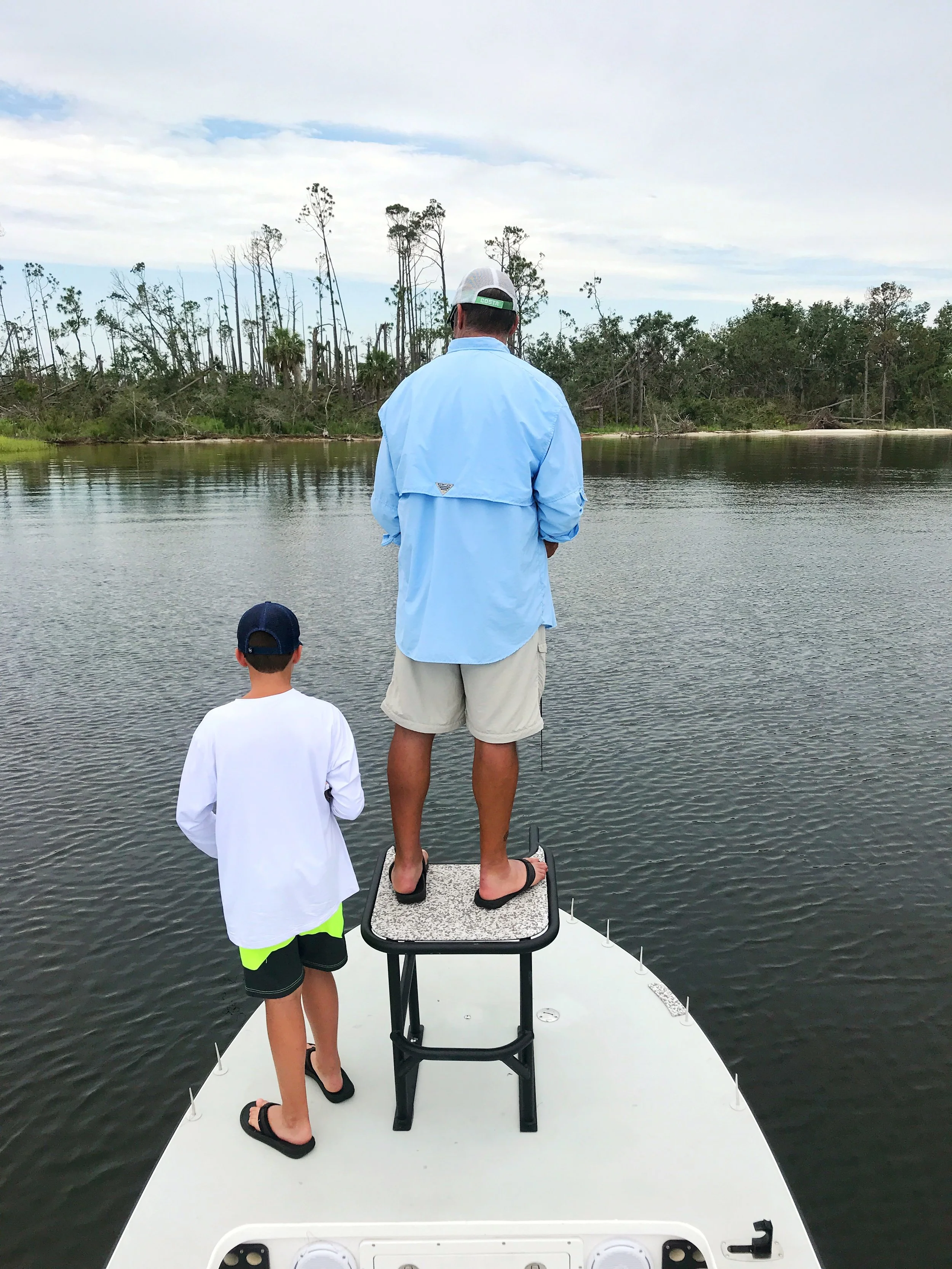 Two boys and a man stand on a boat fishing in a river, surrounded by trees and cloudy sky.