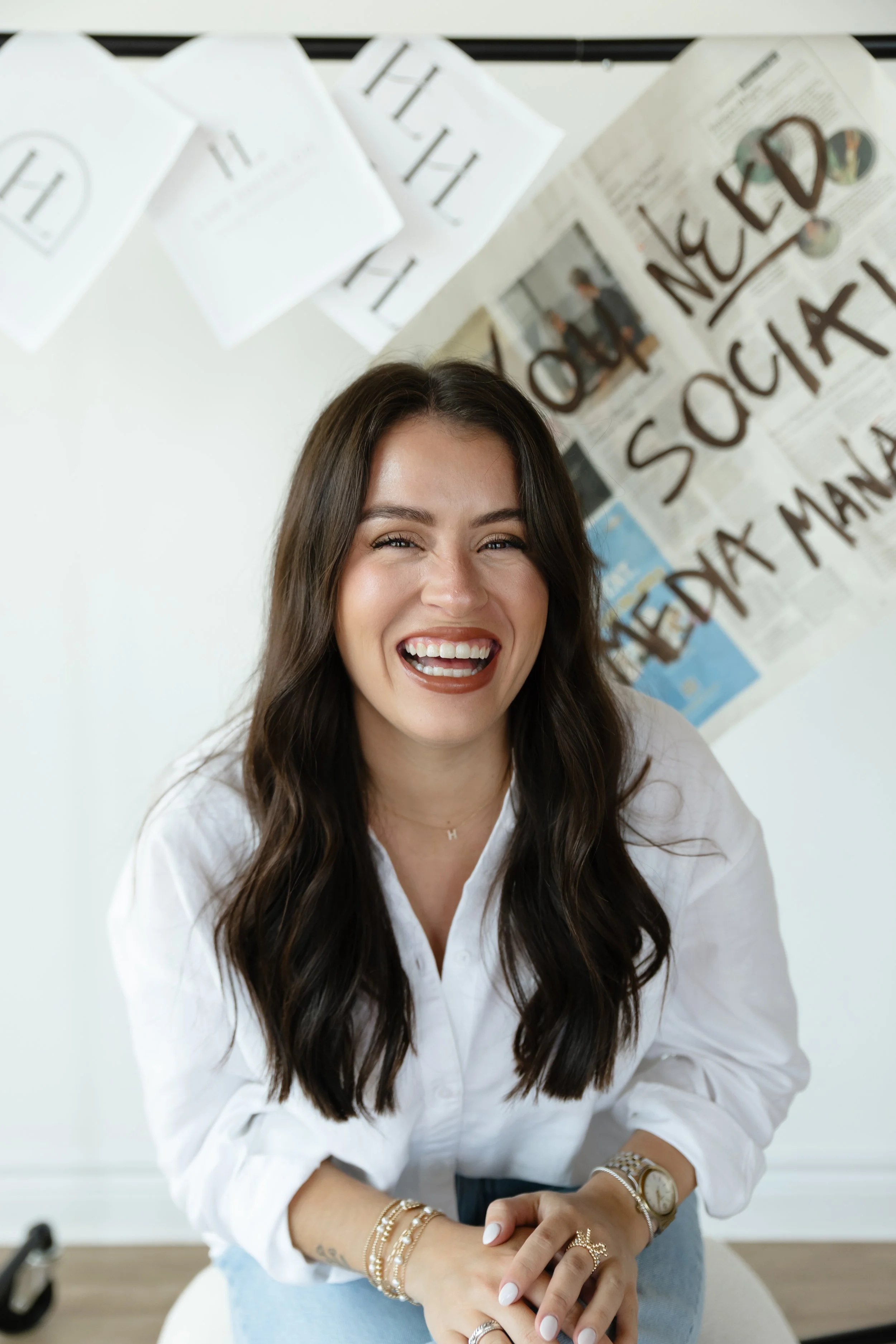 Woman with long dark hair wearing a white shirt, smiling with closed eyes, showing her teeth, seated with her hands folded on her lap, wearing multiple gold bracelets and rings, in a bright room with a wall covered in posters and papers, including one with bold black writing.